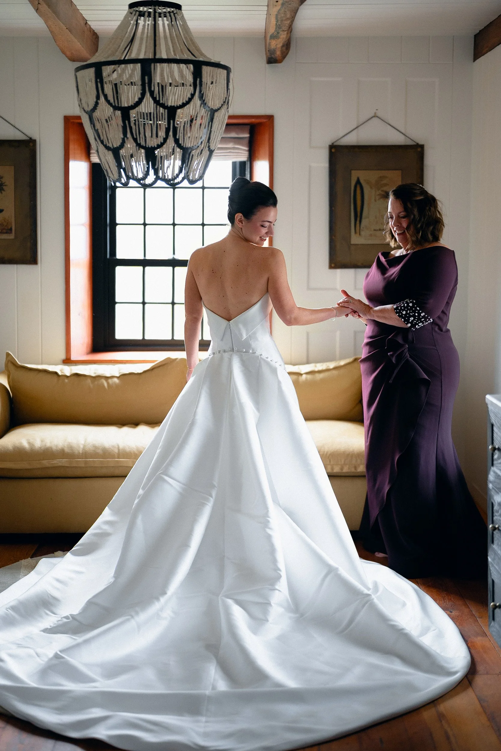A bride in a white wedding dress standing next to a woman in a purple gown. They are holding hands and smiling indoors with a window, paintings, and a large chandelier above them.
