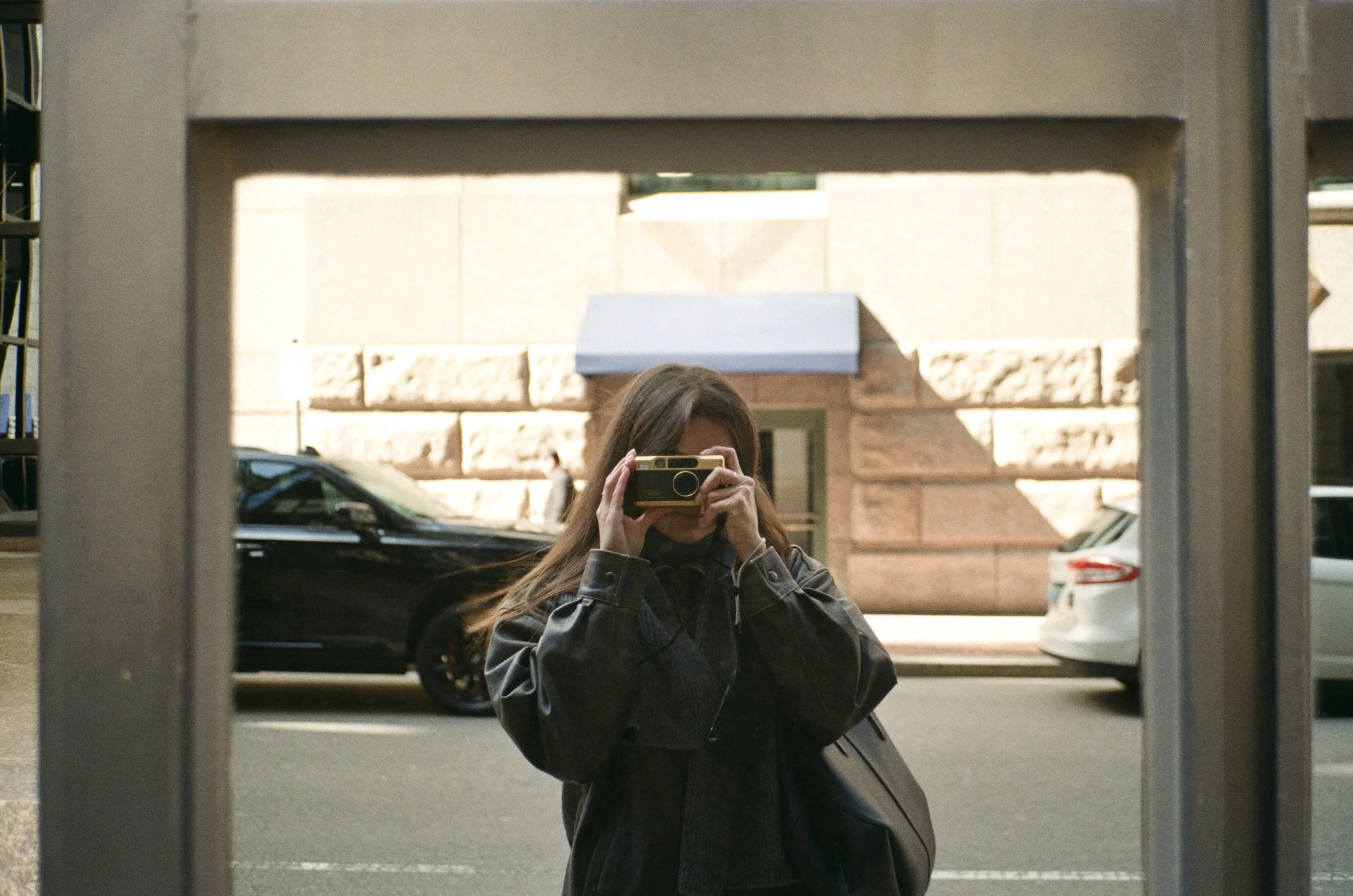A woman taking a photograph of herself with a camera in a mirror on a city sidewalk.
