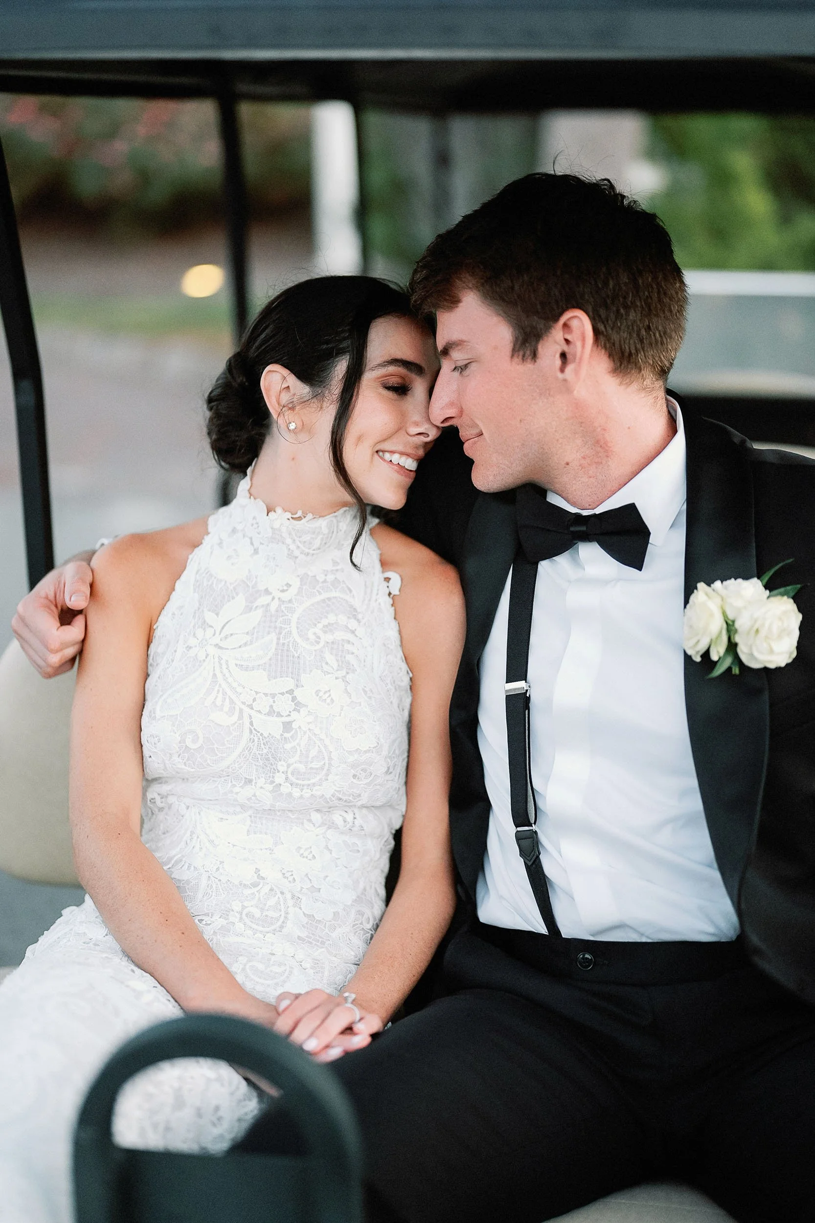 A bride and groom sitting closely together in a golf cart, smiling with their foreheads touching, dressed in wedding attire.