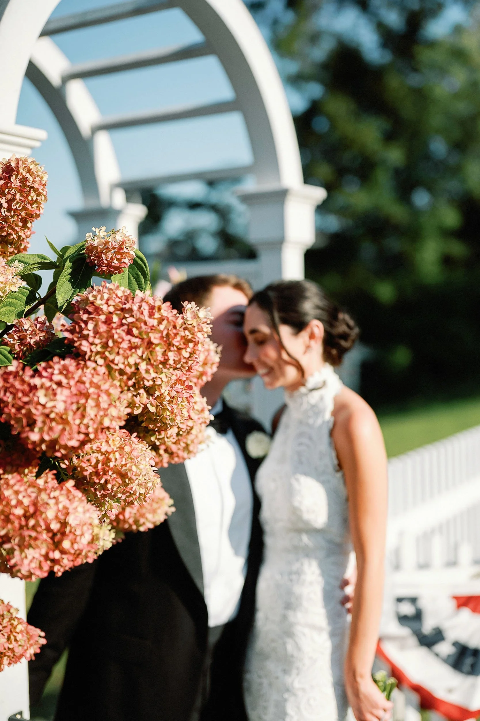 A same-sex couple sharing a kiss on their wedding day, standing outdoors near a white garden arch decorated with pink hydrangea flowers.