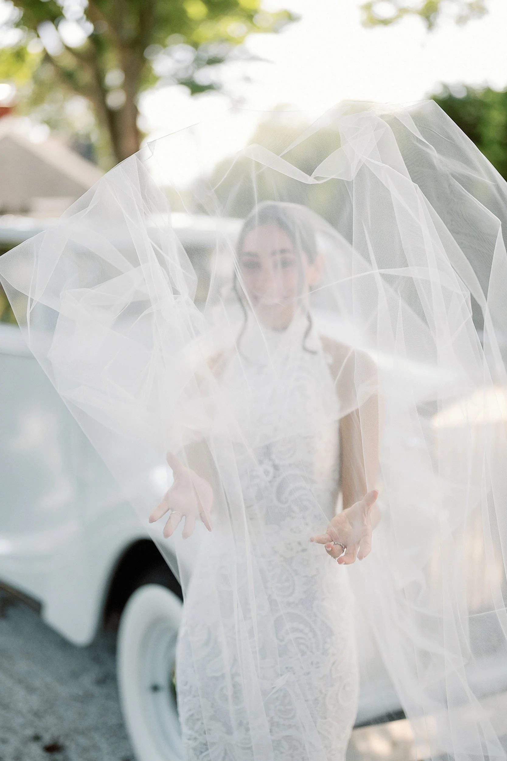 Bride standing outside, looking through veil with hands extended, wearing a white lace wedding dress, with a vintage car in the background.