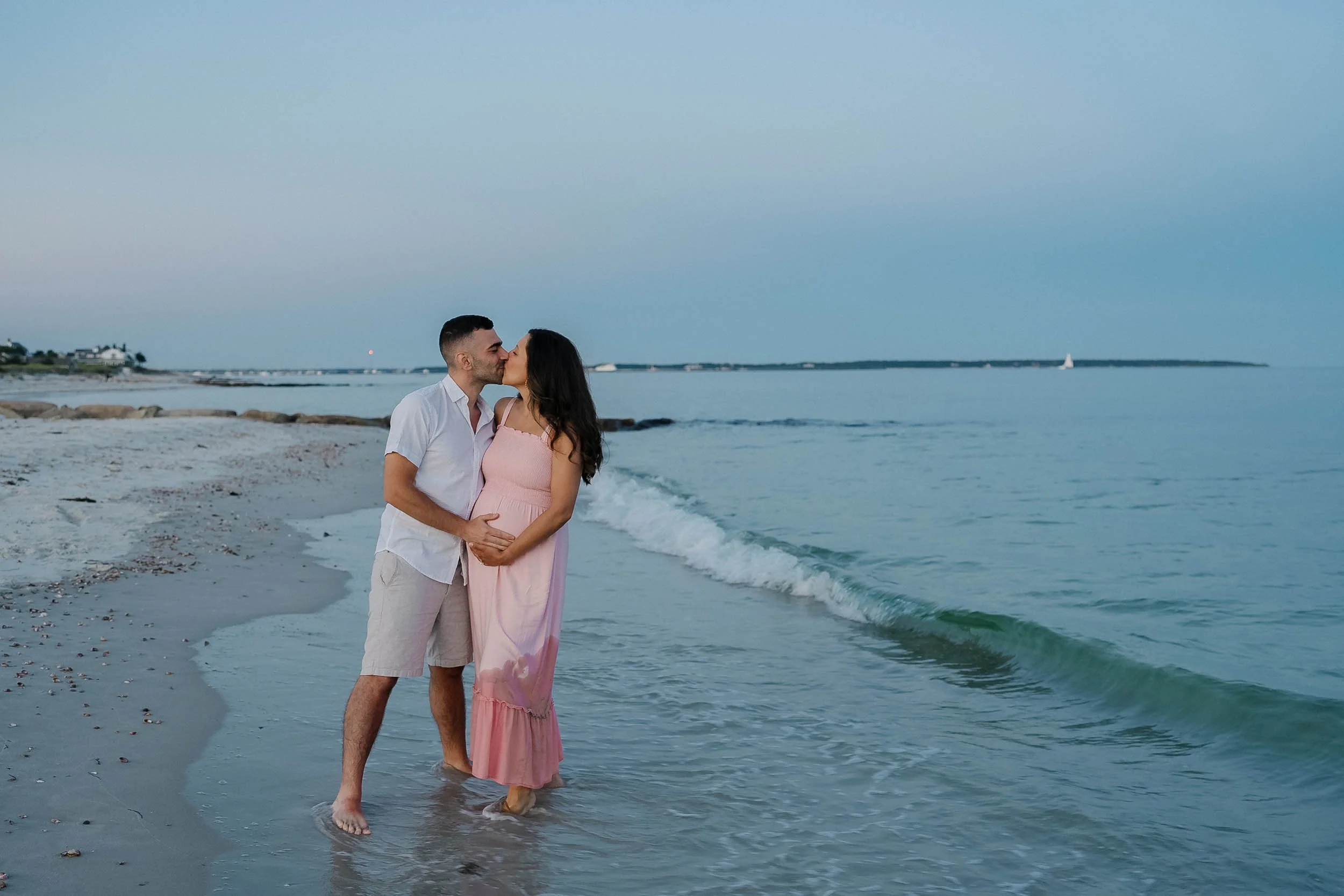 A couple kissing on a beach, standing in shallow water at sunset.