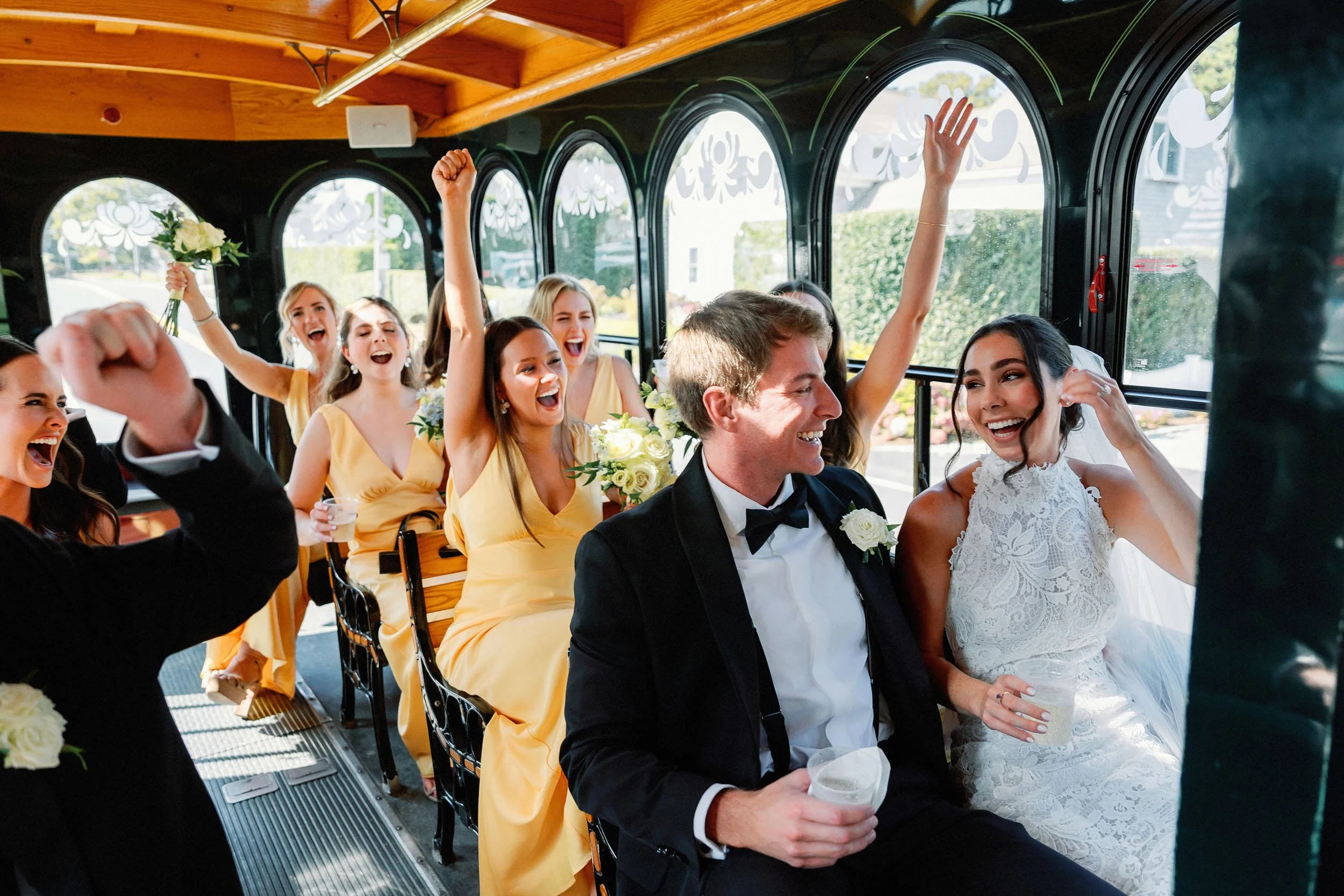 A group of people celebrating on a bus, with a bride and groom sitting at the front, smiling and laughing, surrounded by bridesmaids in yellow dresses.