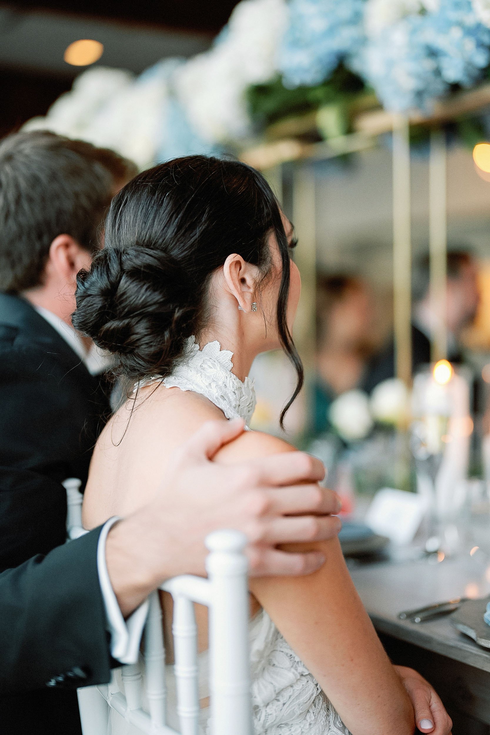 A woman with dark hair styled in an elegant bun and wearing a lace dress, sitting at a table during a formal event.