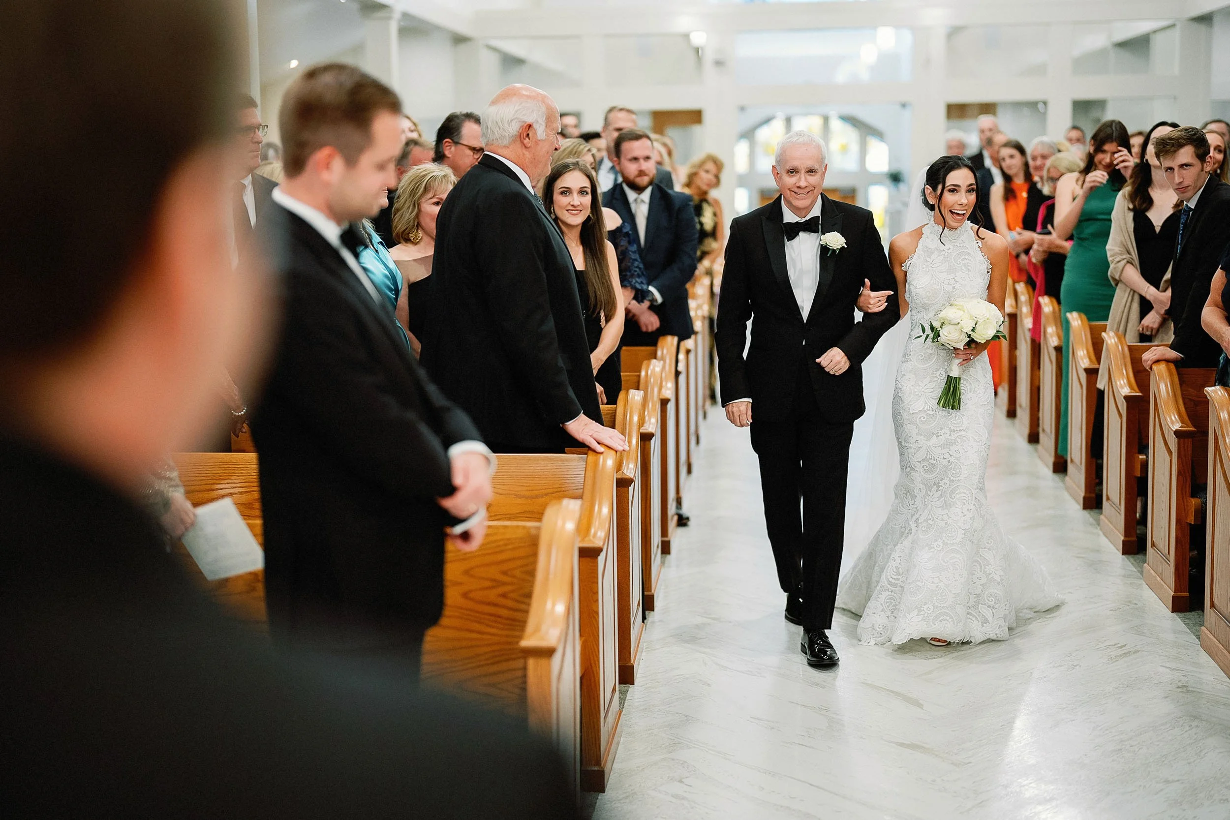 A bride walking down the aisle with her father at a wedding ceremony, surrounded by guests seated in wooden pews inside a brightly lit church.
