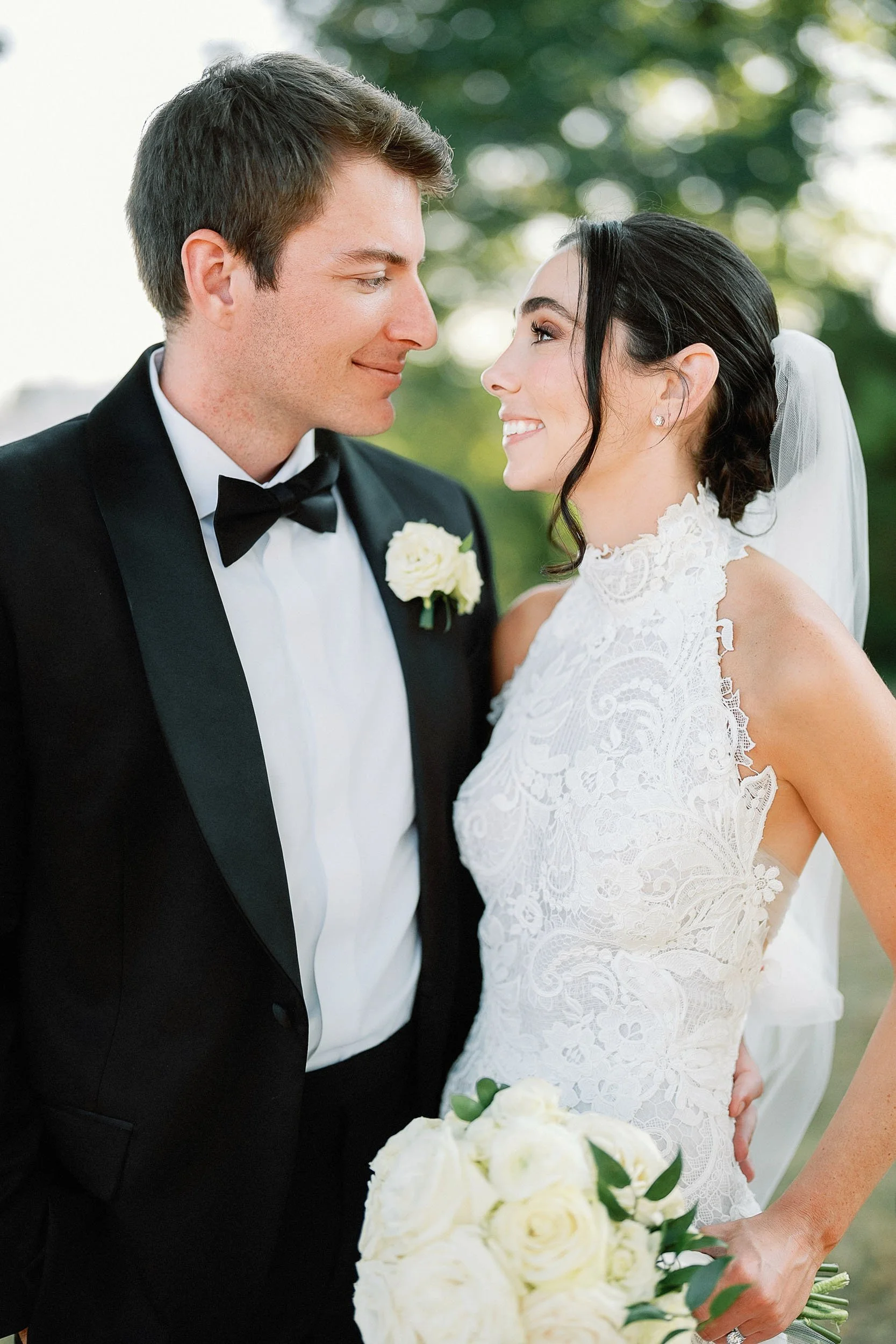 A bride and groom gazing into each other's eyes outdoors, with the groom in a tuxedo and the bride in a lace wedding dress holding a bouquet of white roses.