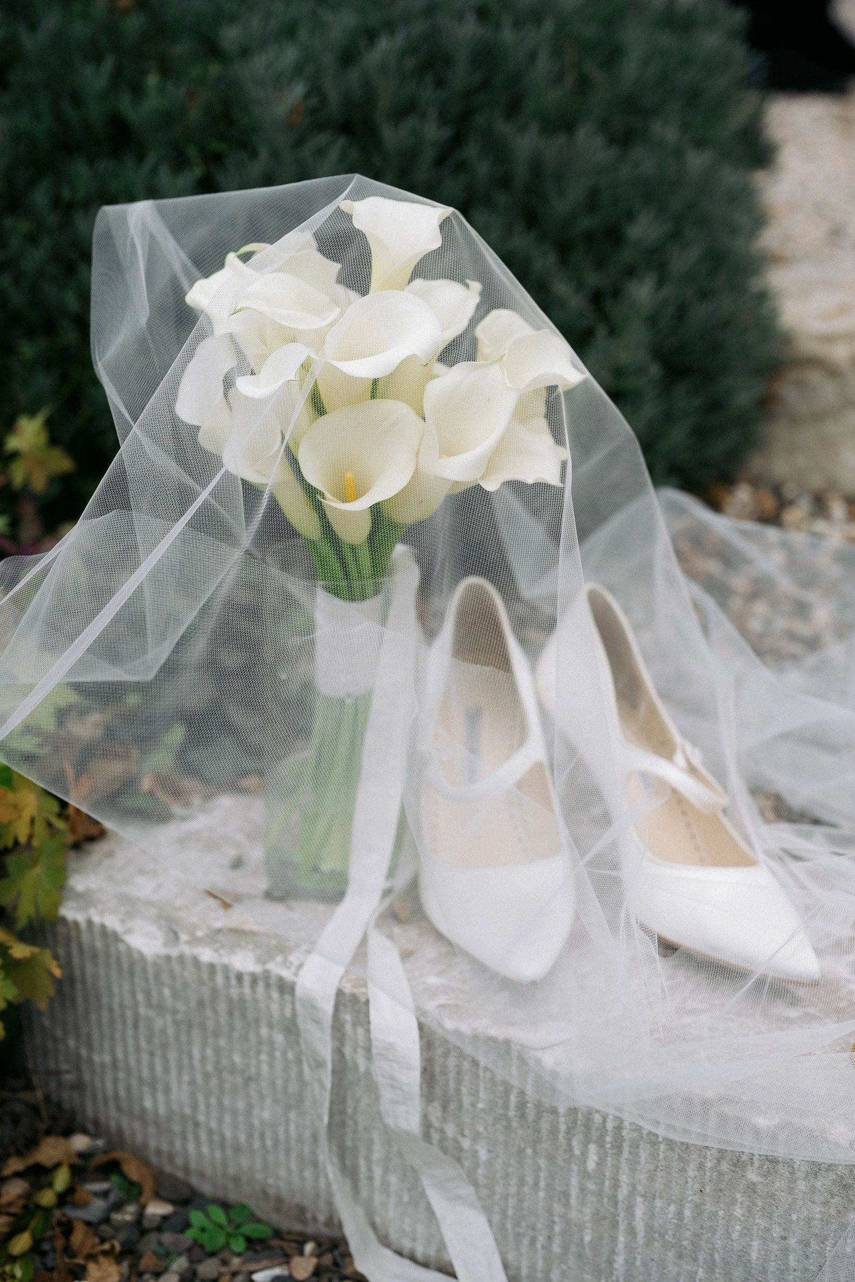 A bouquet of white calla lilies with a veil over white high-heeled shoes, arranged on a stone surface outdoors.