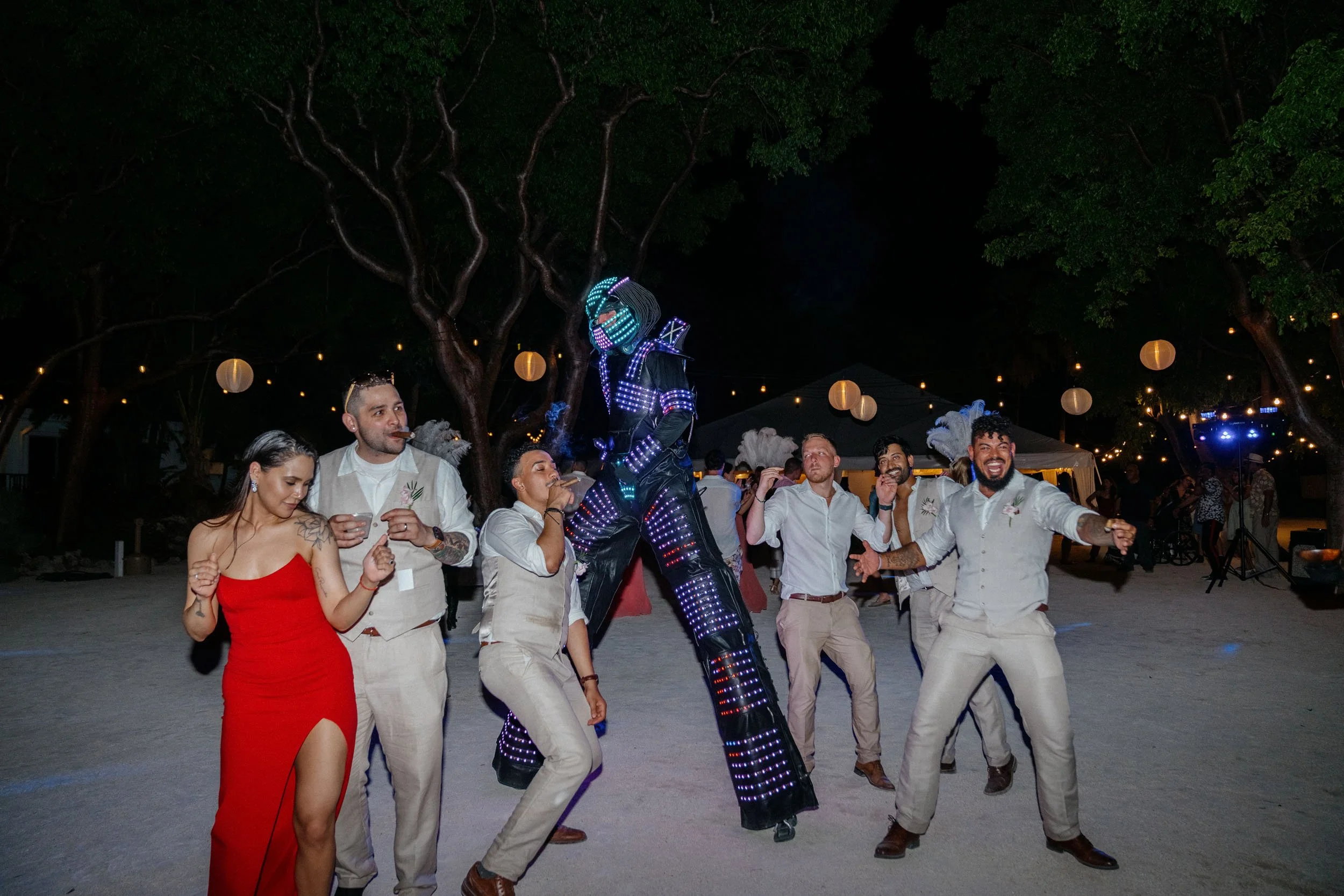 People dancing and celebrating at an outdoor event during the night with trees, string lights, and paper lanterns in the background. A performer dressed in a black, lit-up costume is in the center.