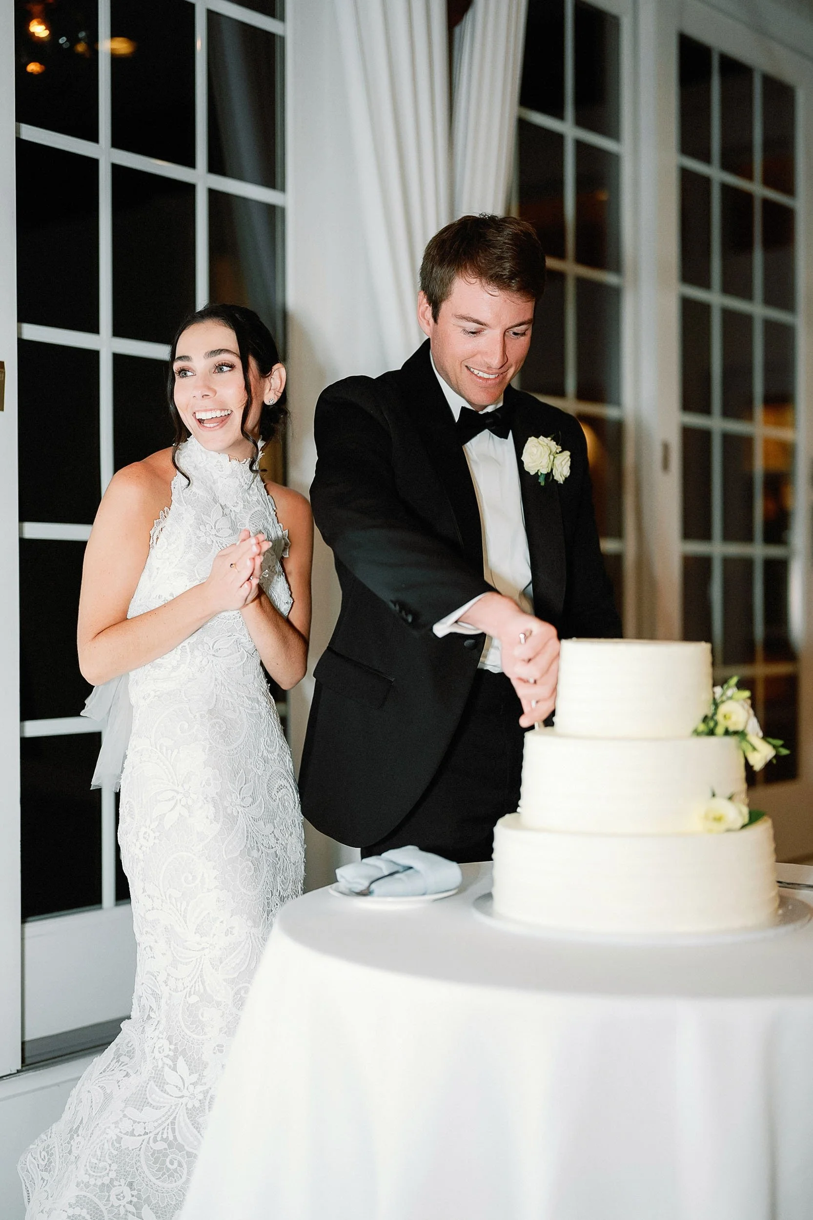 A newlywed couple cuts a wedding cake together. The bride is wearing a white lace wedding dress, and the groom is wearing a black tuxedo with a white shirt and black bow tie.