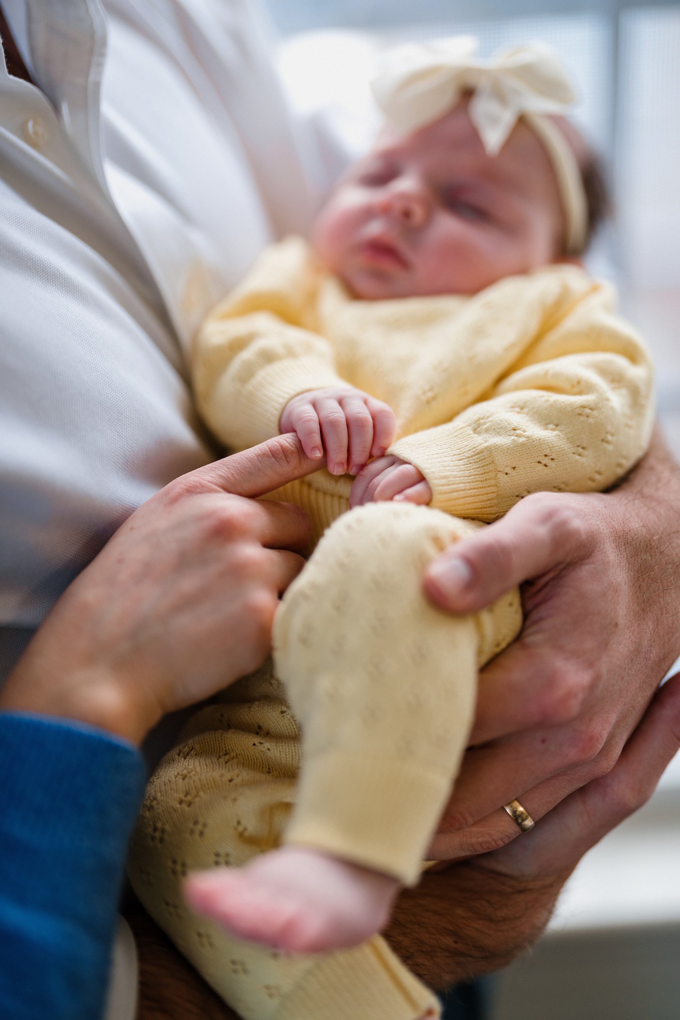 Close-up of a newborn baby in yellow clothing and a bow headband, being held and gently grasped by an adult, with another adult's hand touching the baby's foot.