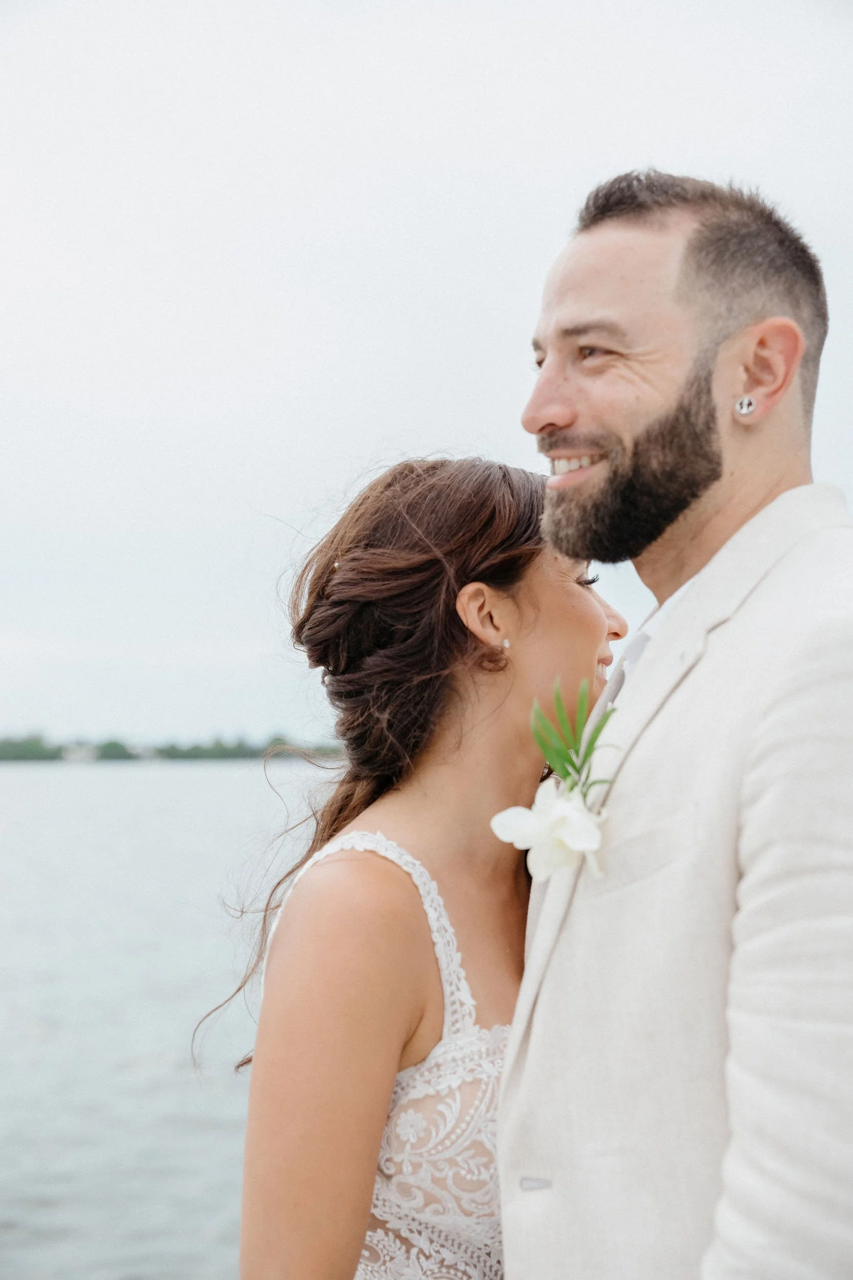 A bride and groom standing close to each other outdoors near water, with the bride resting her head on the groom's shoulder, both smiling gently.