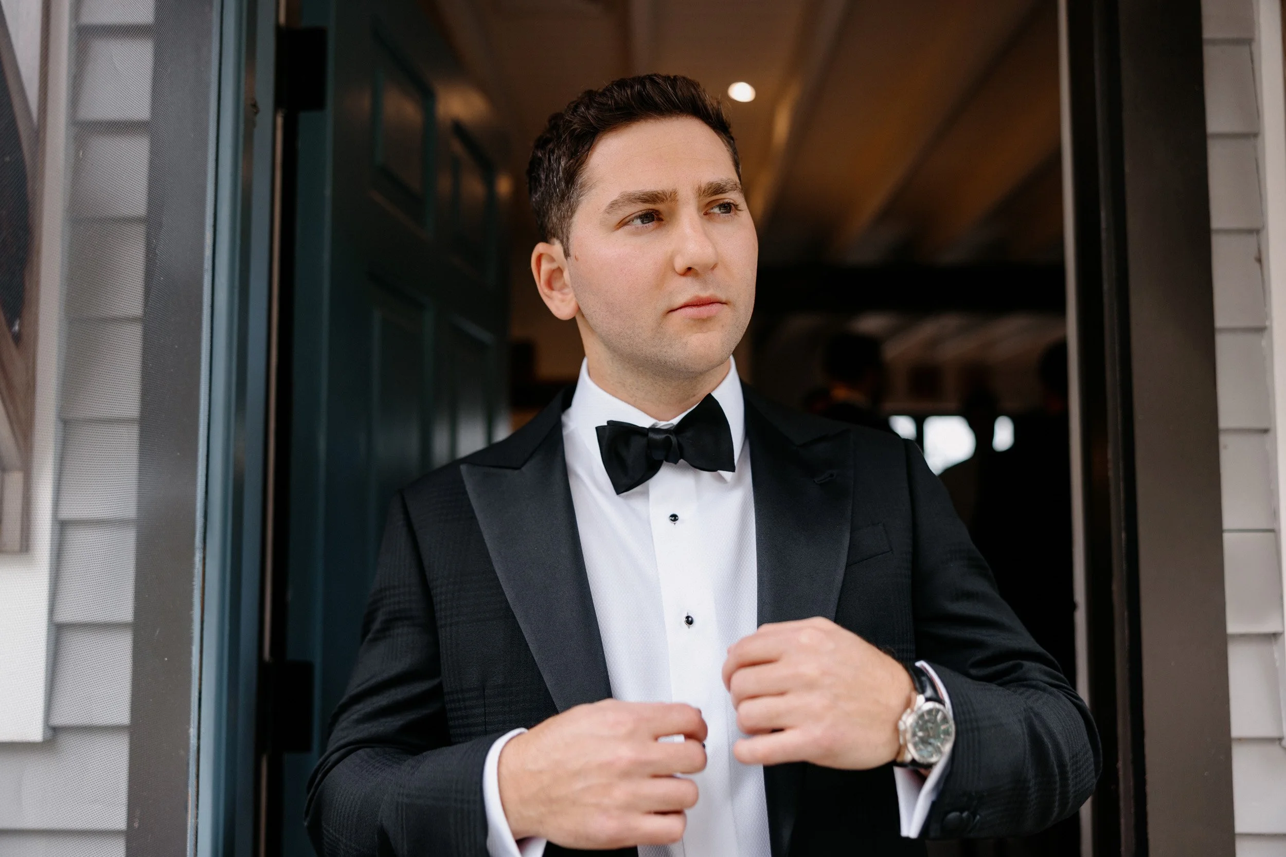 A young man in a tuxedo with a bow tie, adjusting his jacket in front of a house.