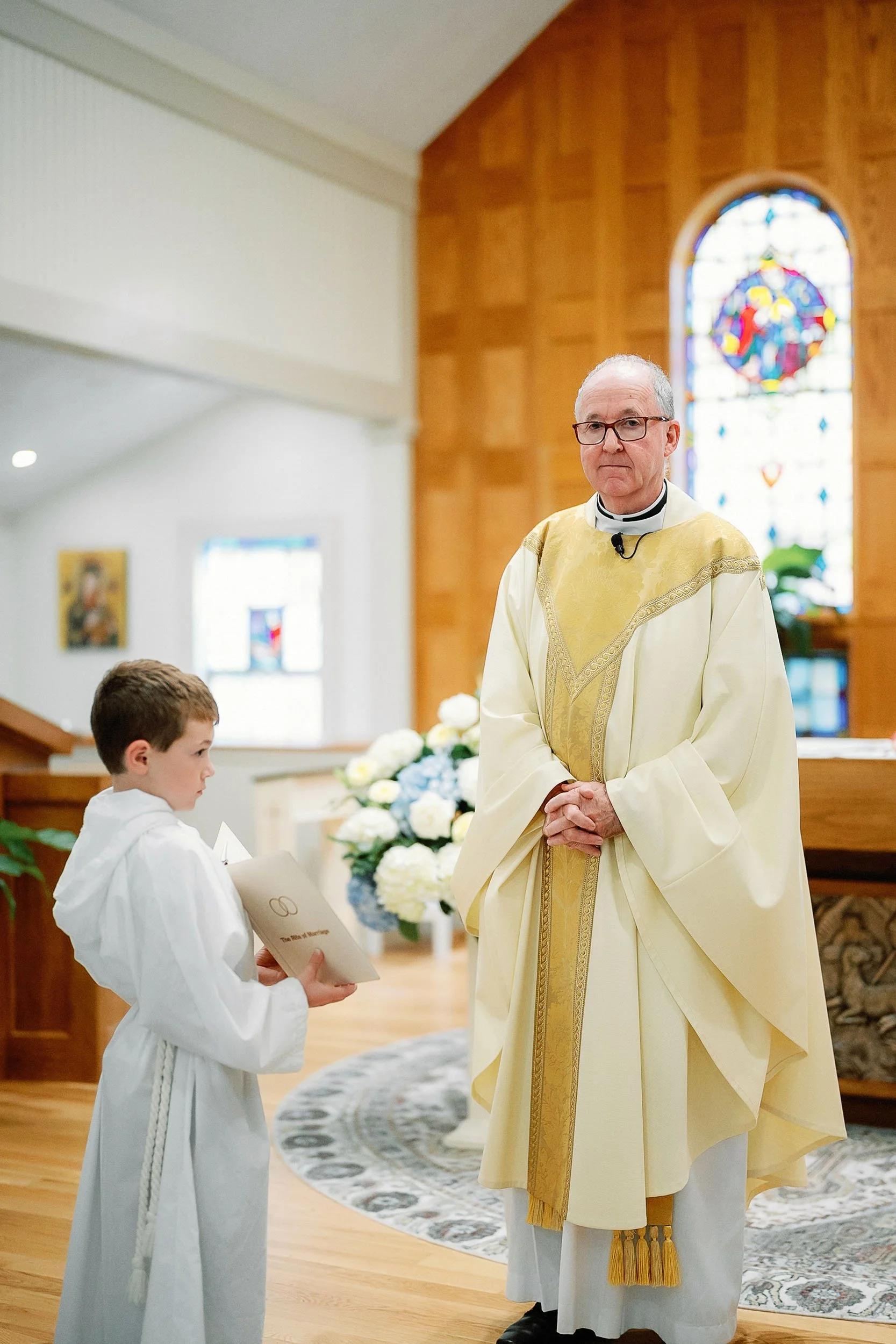 A priest and a young altar server are inside a church during a ceremony. The priest is wearing cream and gold vestments, standing with his hands clasped. The altar server, dressed in white robes, is holding a booklet and looking at the priest. There 