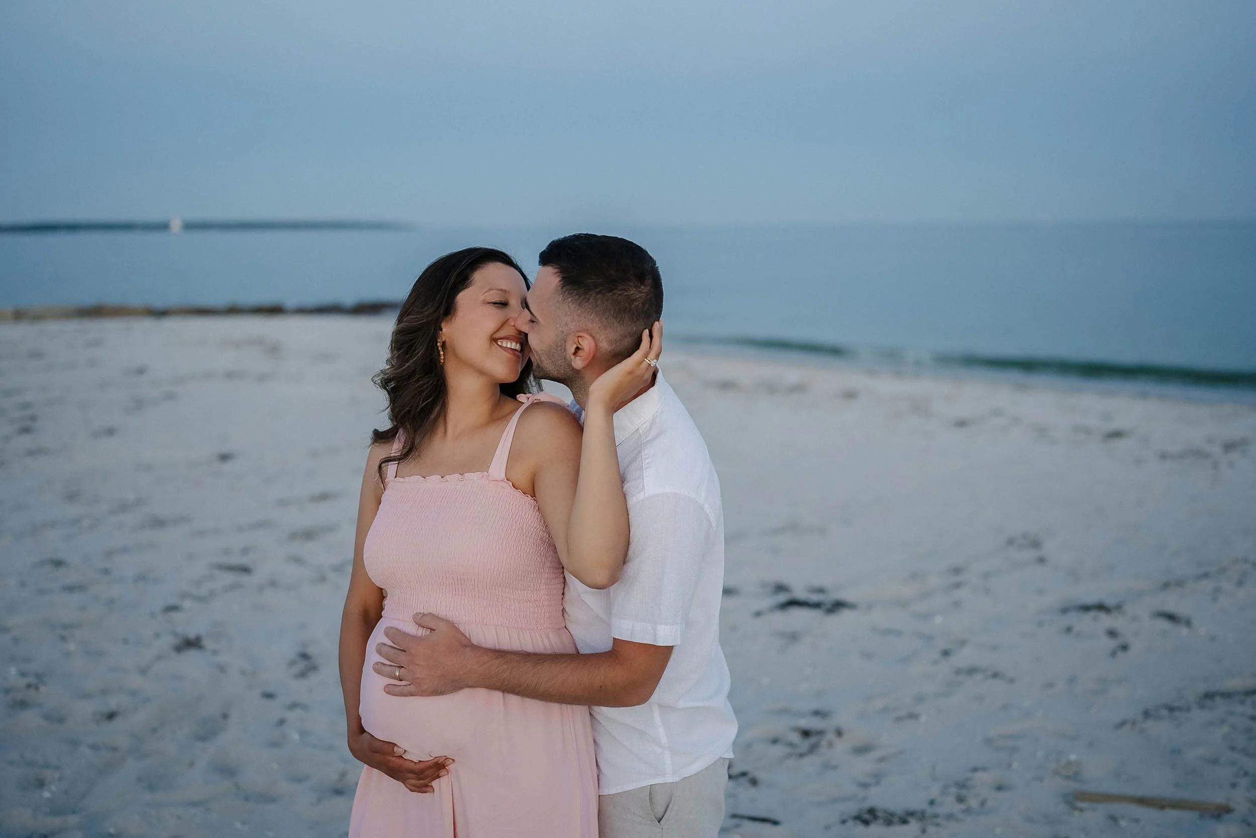 A pregnant woman and a man are embracing and smiling at each other on a beach during sunset.
