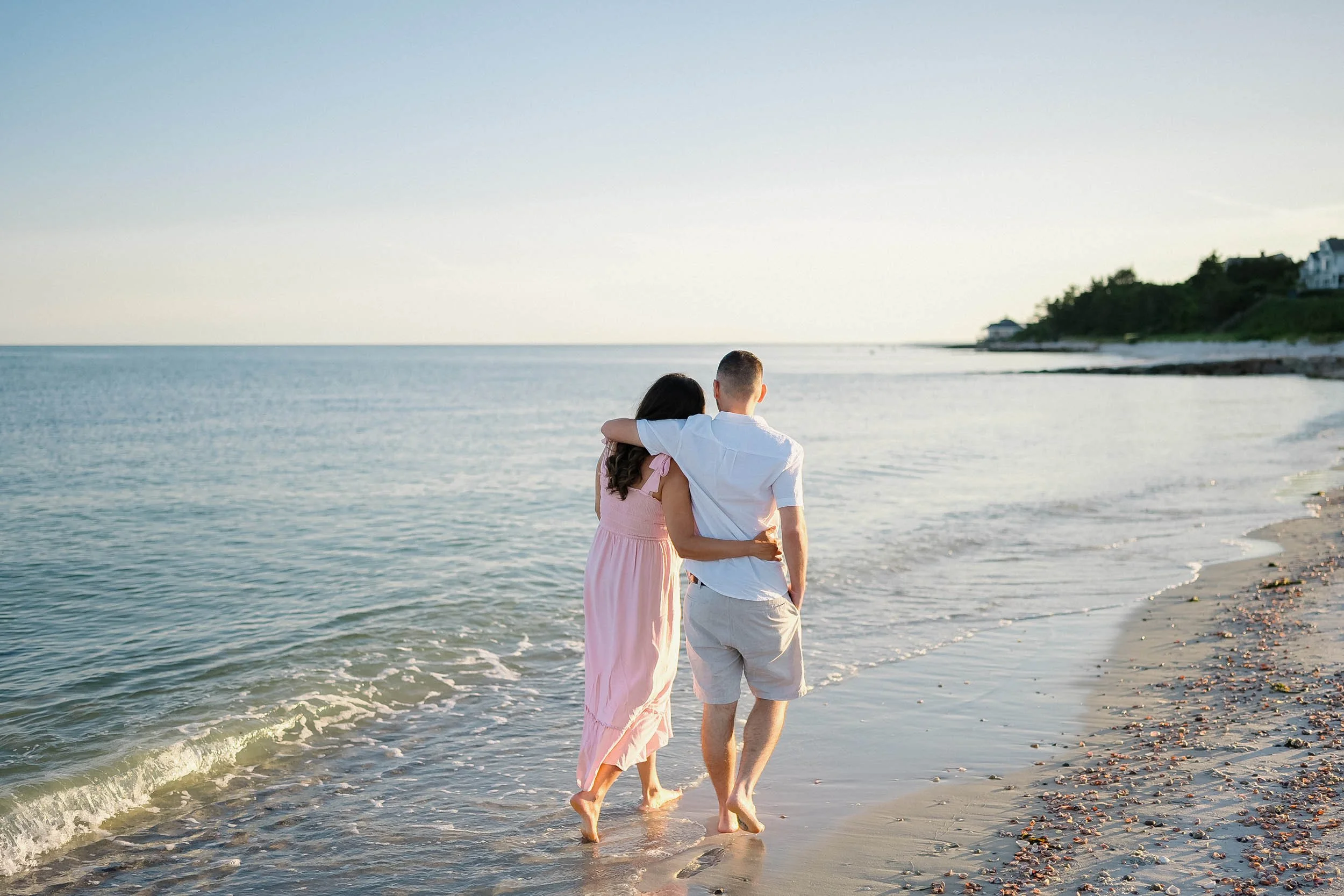 A couple walking on the beach at sunset, with the woman in a pink dress and the man in a white shirt and beige shorts, embracing as they walk along the shoreline.