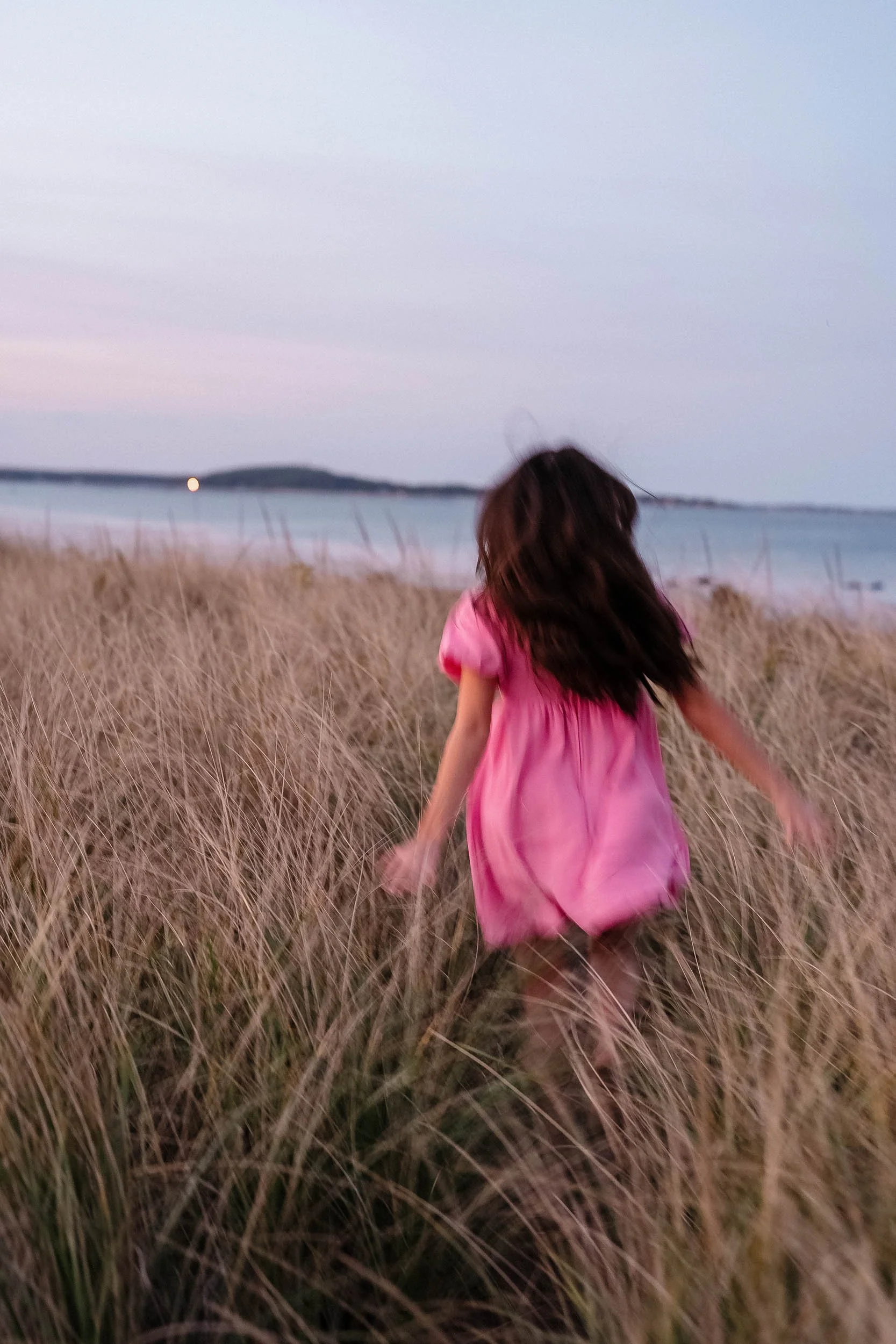 A girl in a pink dress running through tall grass on a beach at dusk.