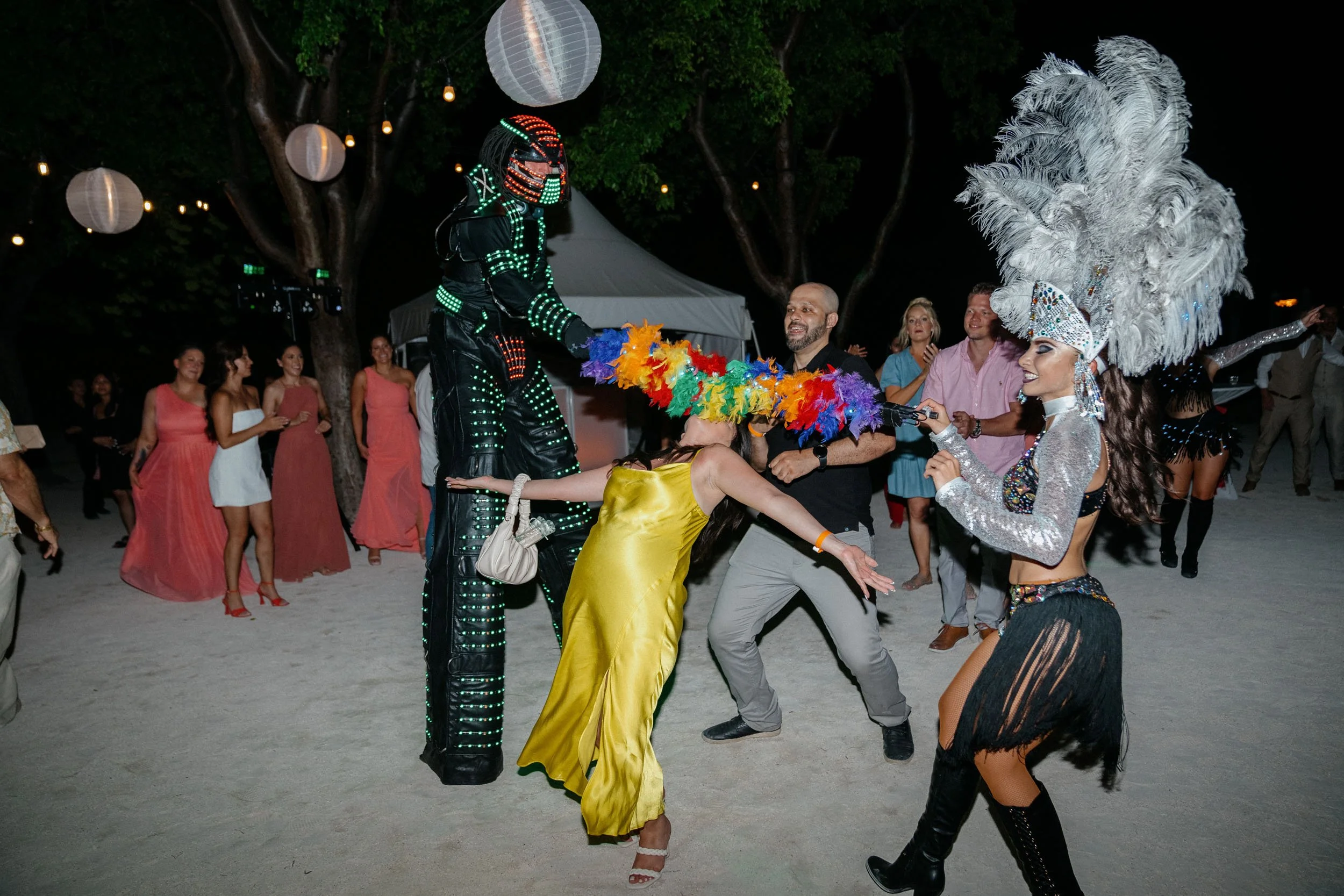 A lively outdoor celebration at night with people dancing and a performer on stilts dressed in a black LED-lit costume, with two women wearing elaborate feathered headdresses. The scene is decorated with paper lanterns hanging from trees.