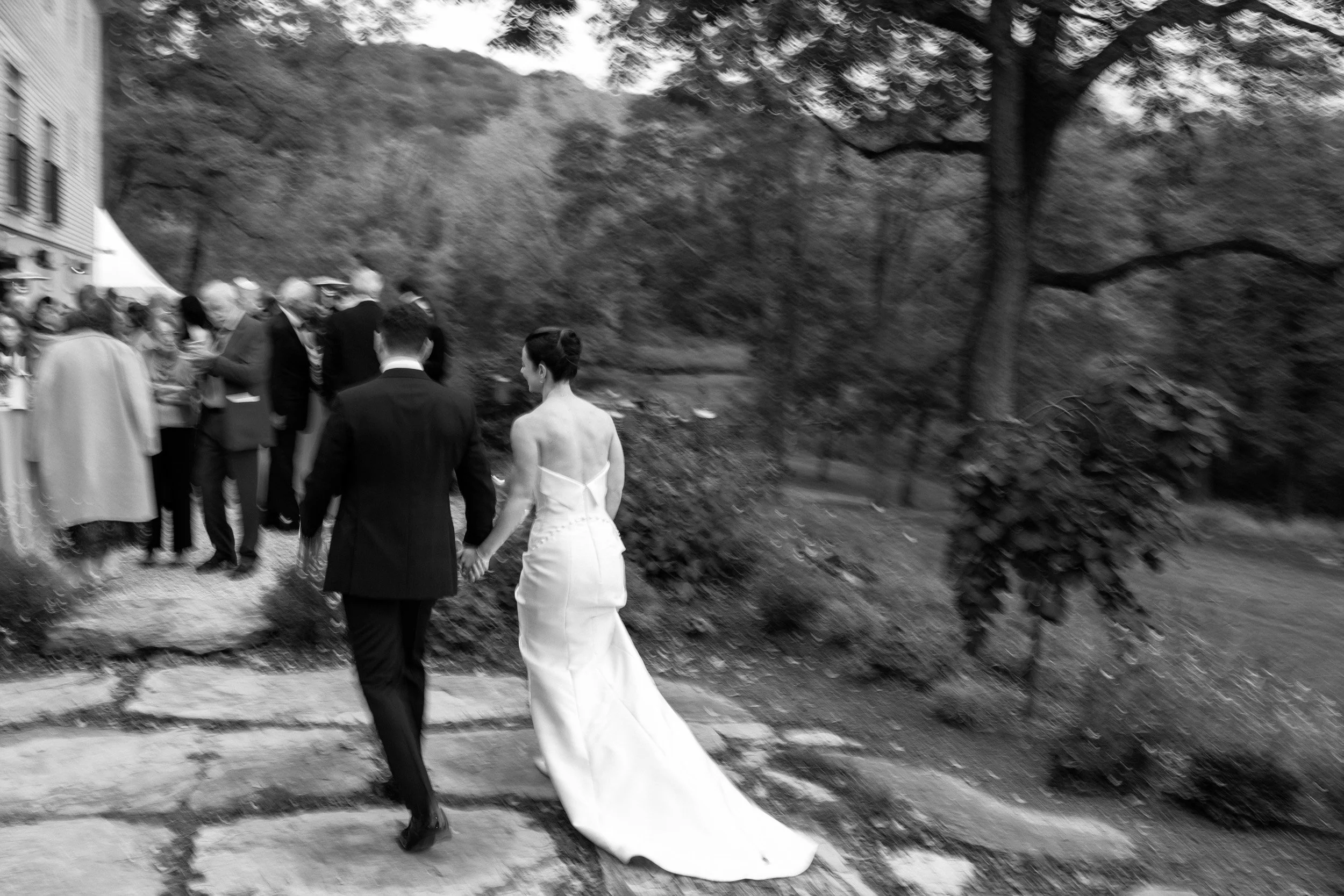 A black and white photo of a bride and groom holding hands, walking on a stone path during a wedding ceremony, with guests gathered near a building and trees in the background.