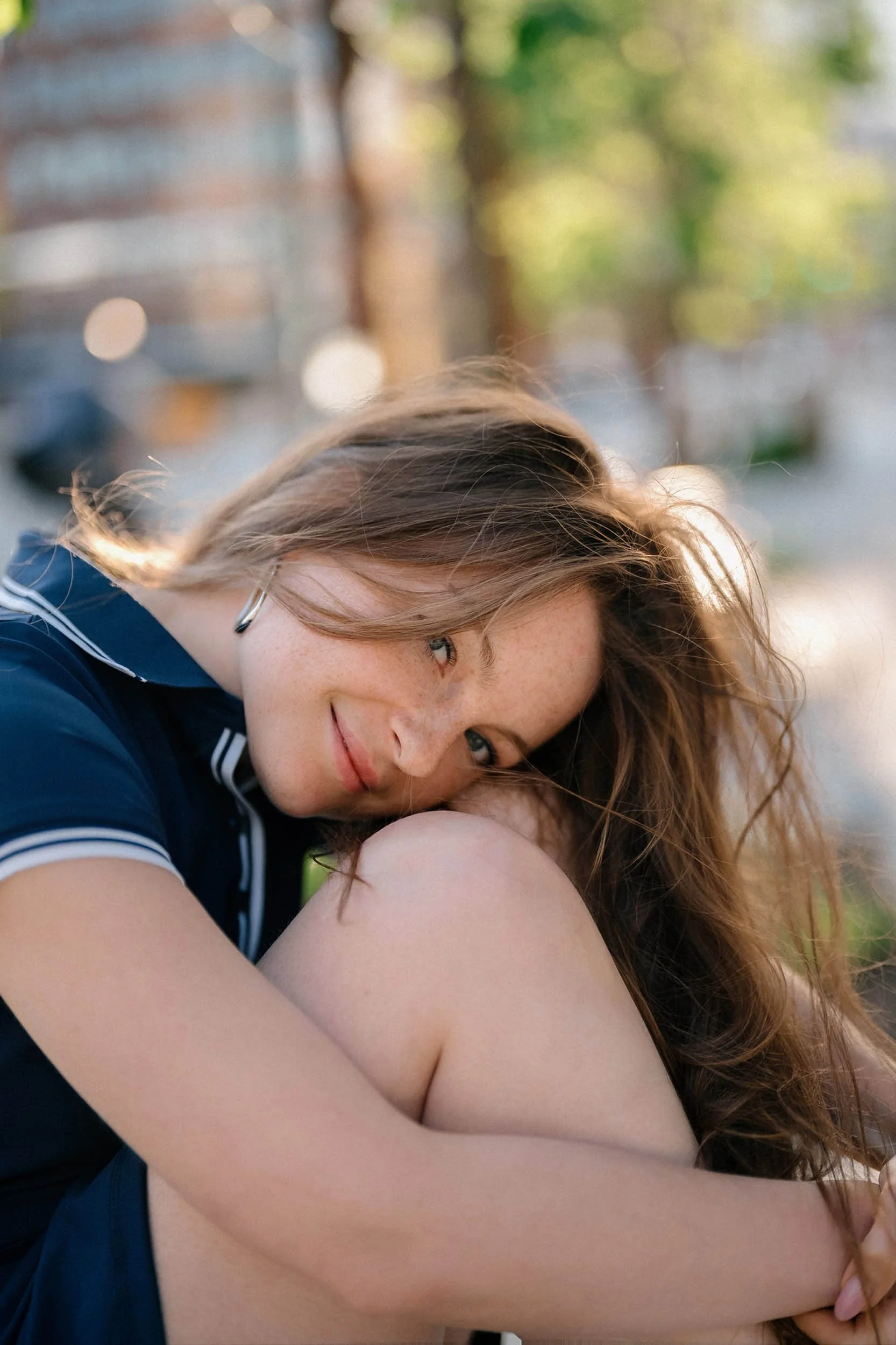 A young woman with long, wavy hair sitting outside during daytime, smiling softly at the camera with her head resting on her knee.