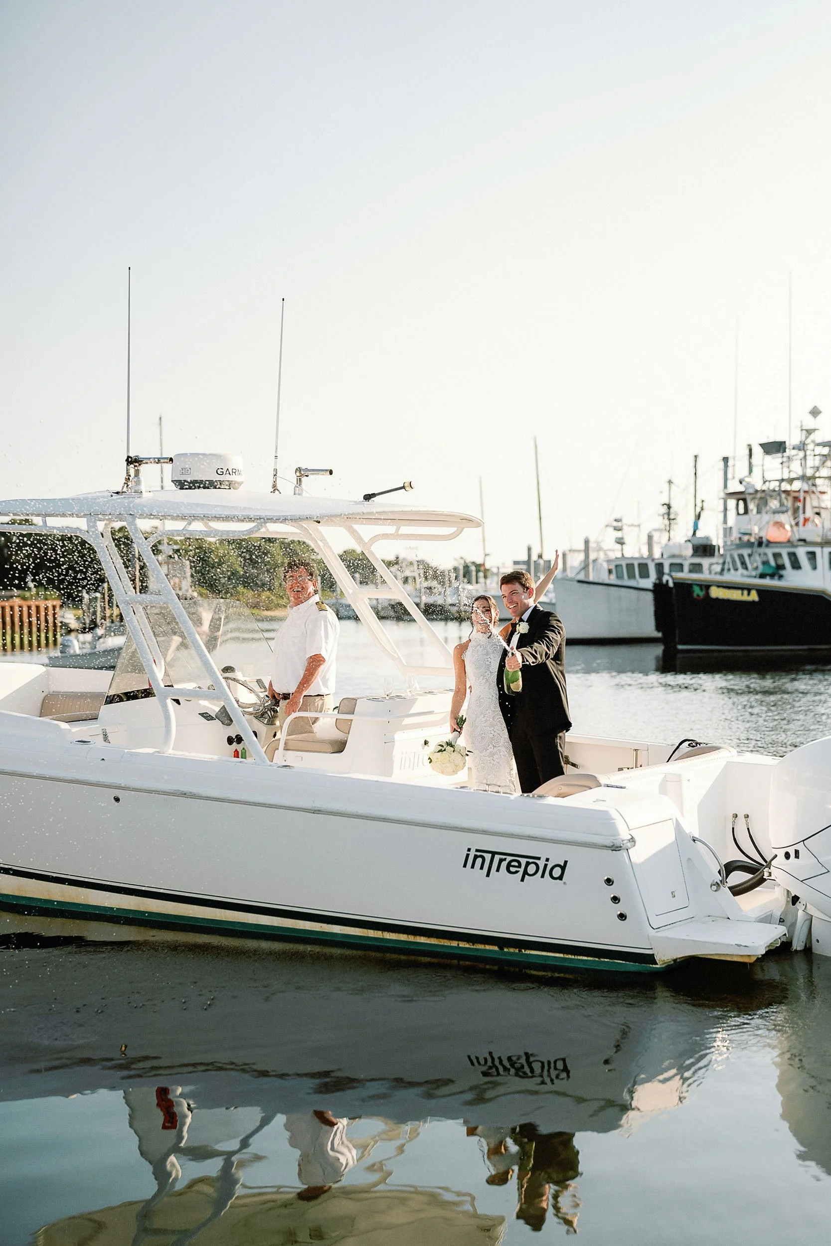 A newlywed couple on a white boat celebrating, with a man steering the boat and a person in white behind the wheel, in a marina with multiple boats docked in the background.