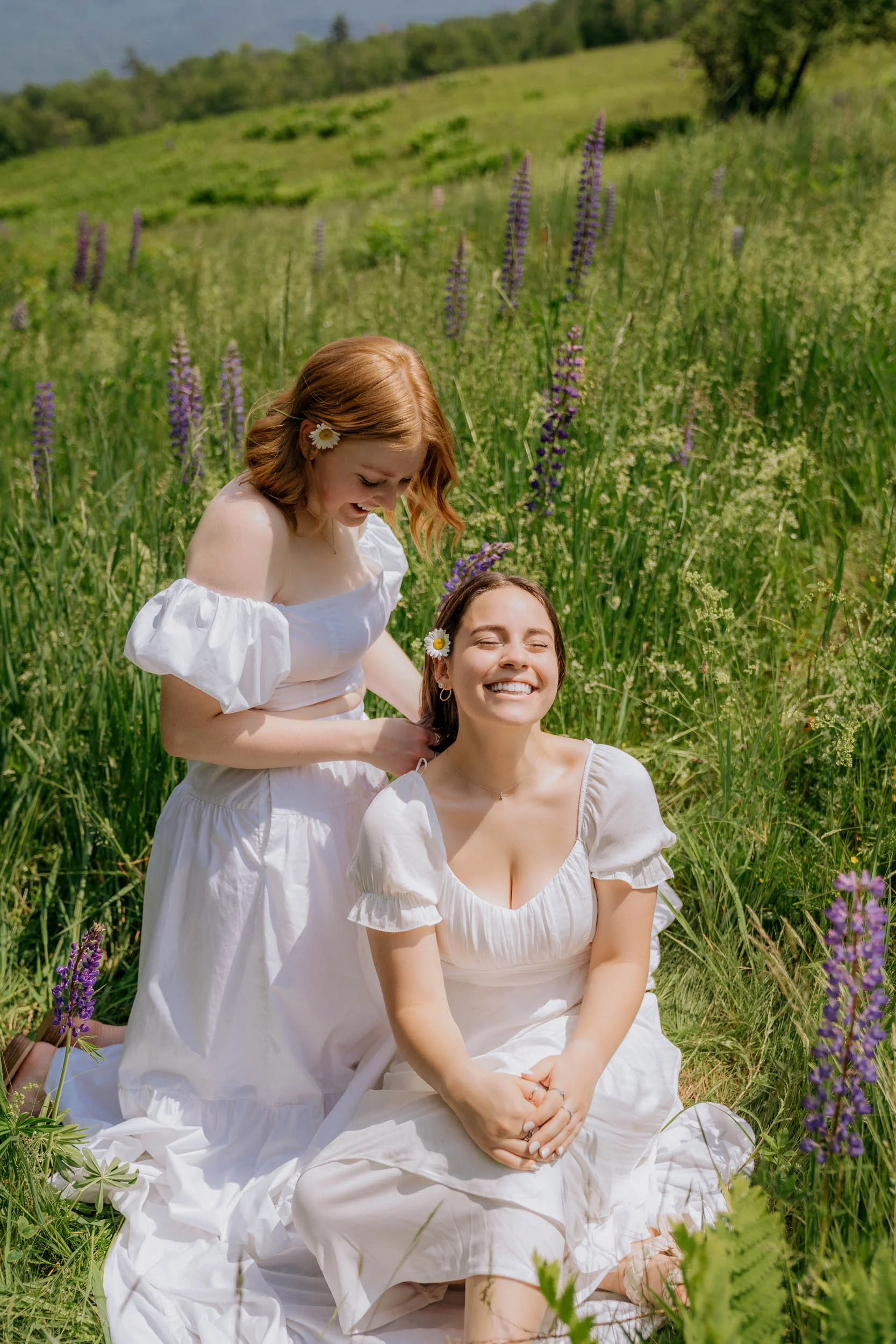Two women in white dresses sitting and kneeling in a field of green grass and purple flowers, smiling and enjoying a sunny day.
