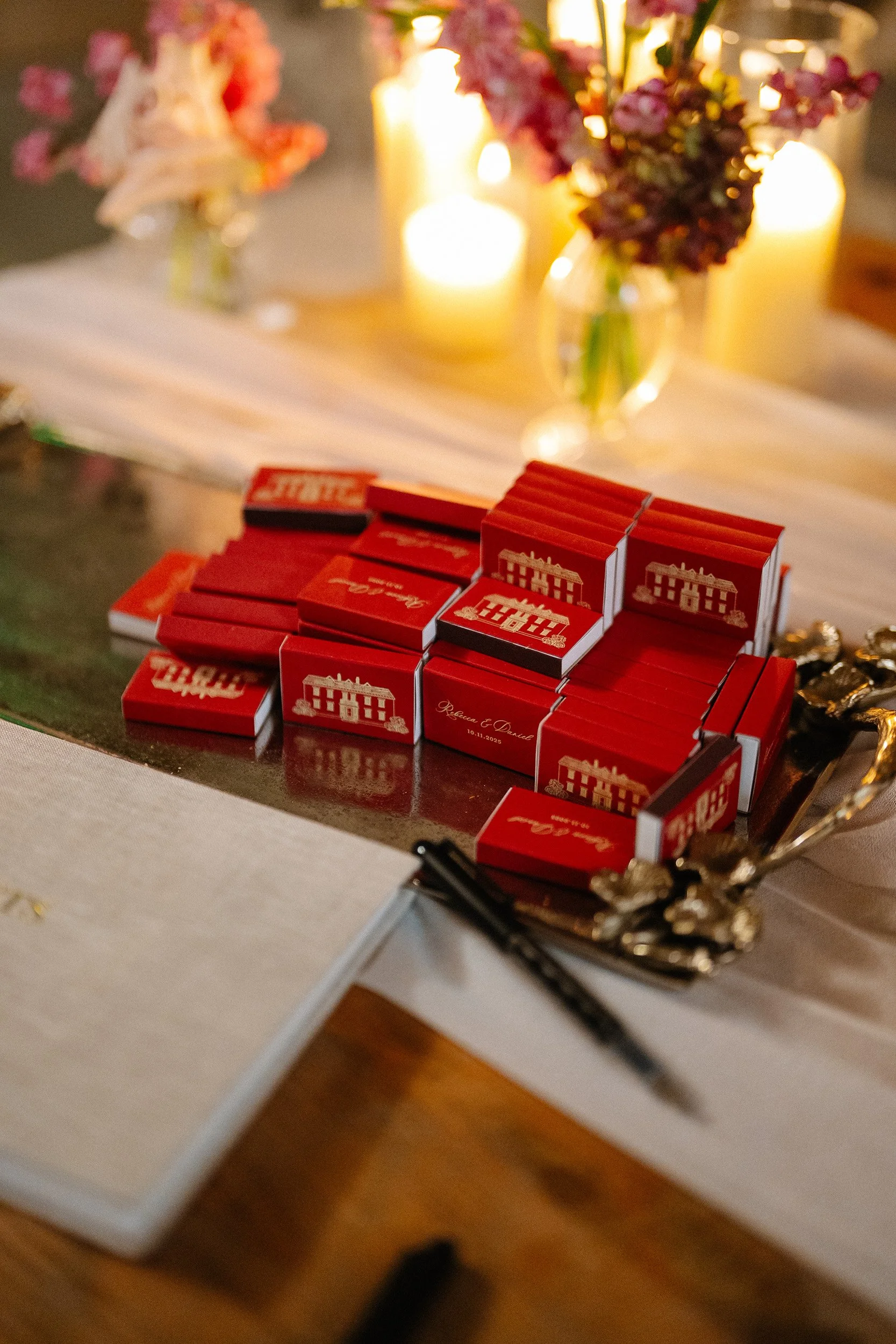 A table with a decorative table runner, lit candles, and vases of pink and purple flowers. In the foreground, there are red matchboxes with white illustrations of a building and the names "Rebecca & Daniel" and a date, along with some pens and jewelr