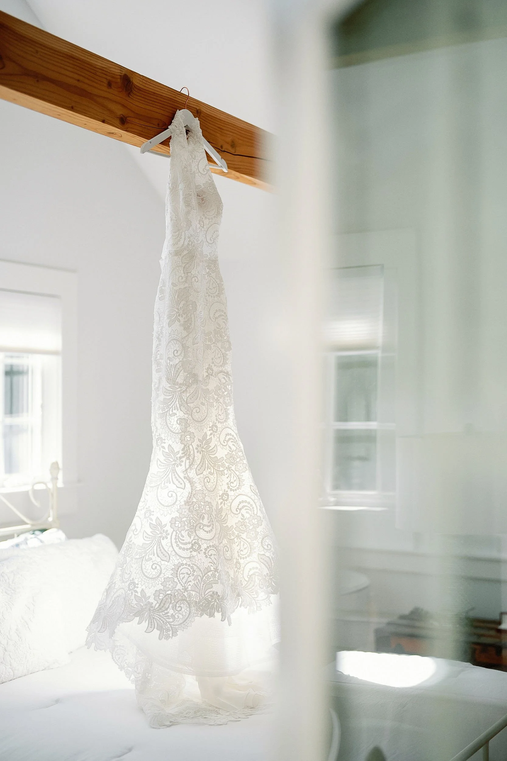 A white lace wedding dress hanging on a wooden beam in a bright room.