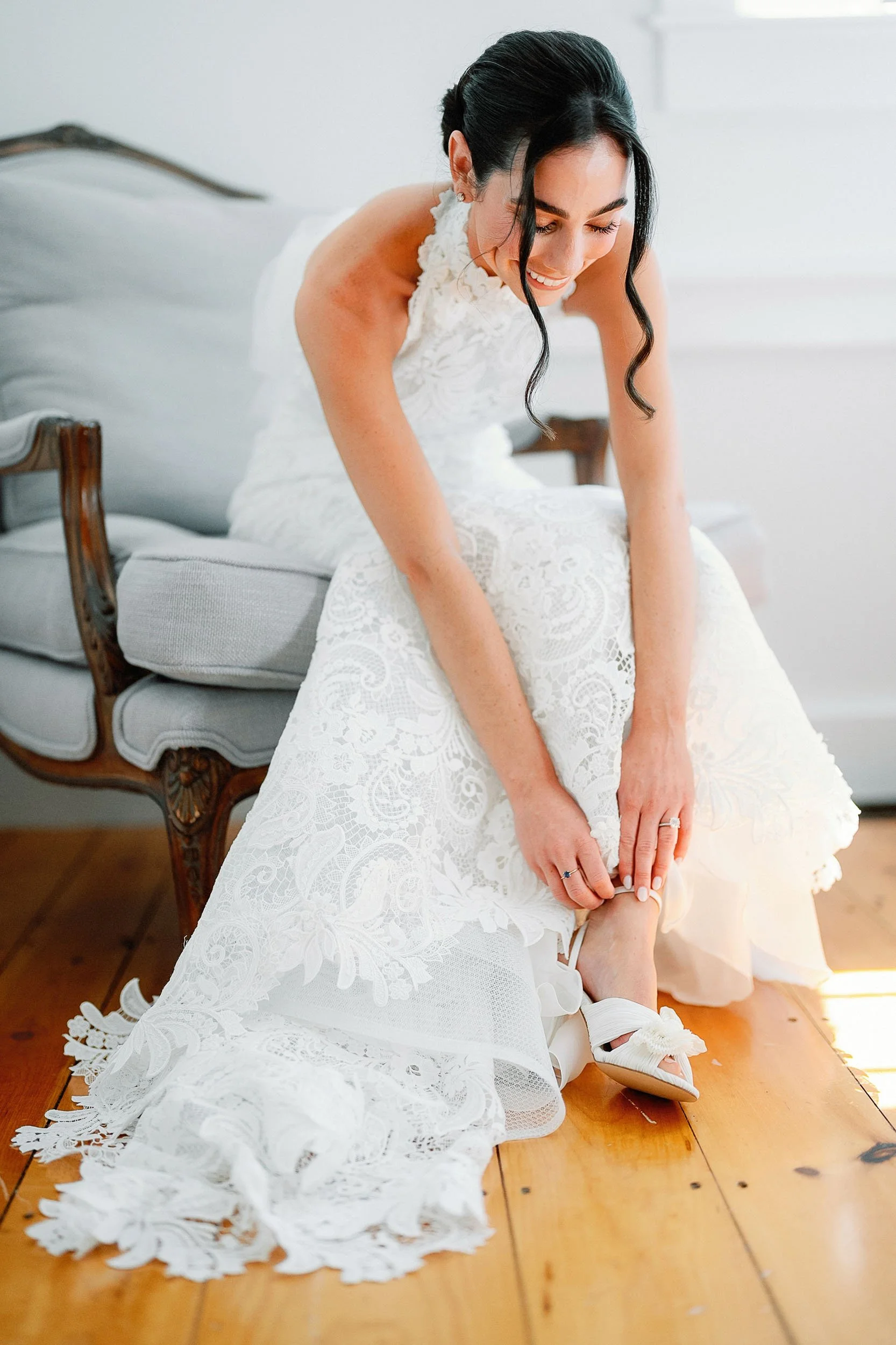 A bride in a white lace wedding dress sitting on a wooden floor, adjusting her white high-heeled sandal.