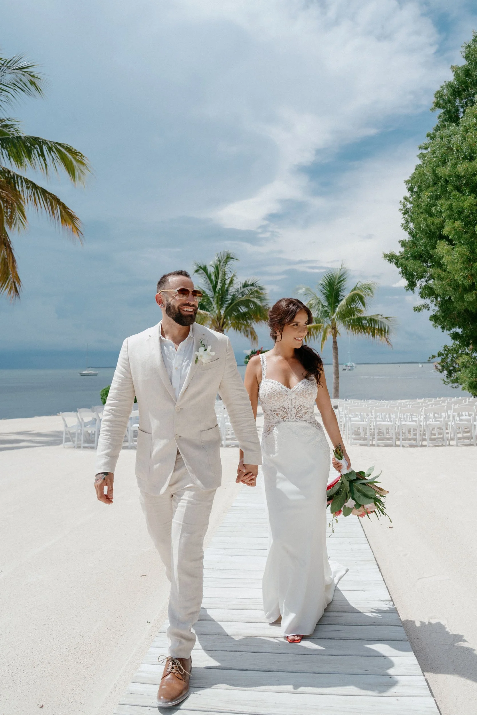 A bride and groom walking hand in hand on a beach wedding ceremony setup with chairs, palm trees, and ocean in the background.
