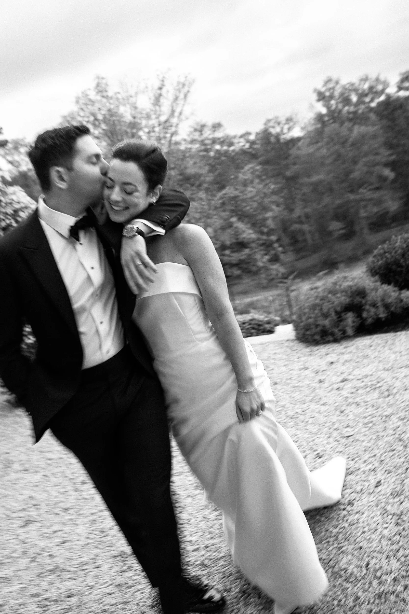 A black and white photo of a happy couple dressed in formal attire, embracing outdoors on a gravel path with trees and bushes in the background.