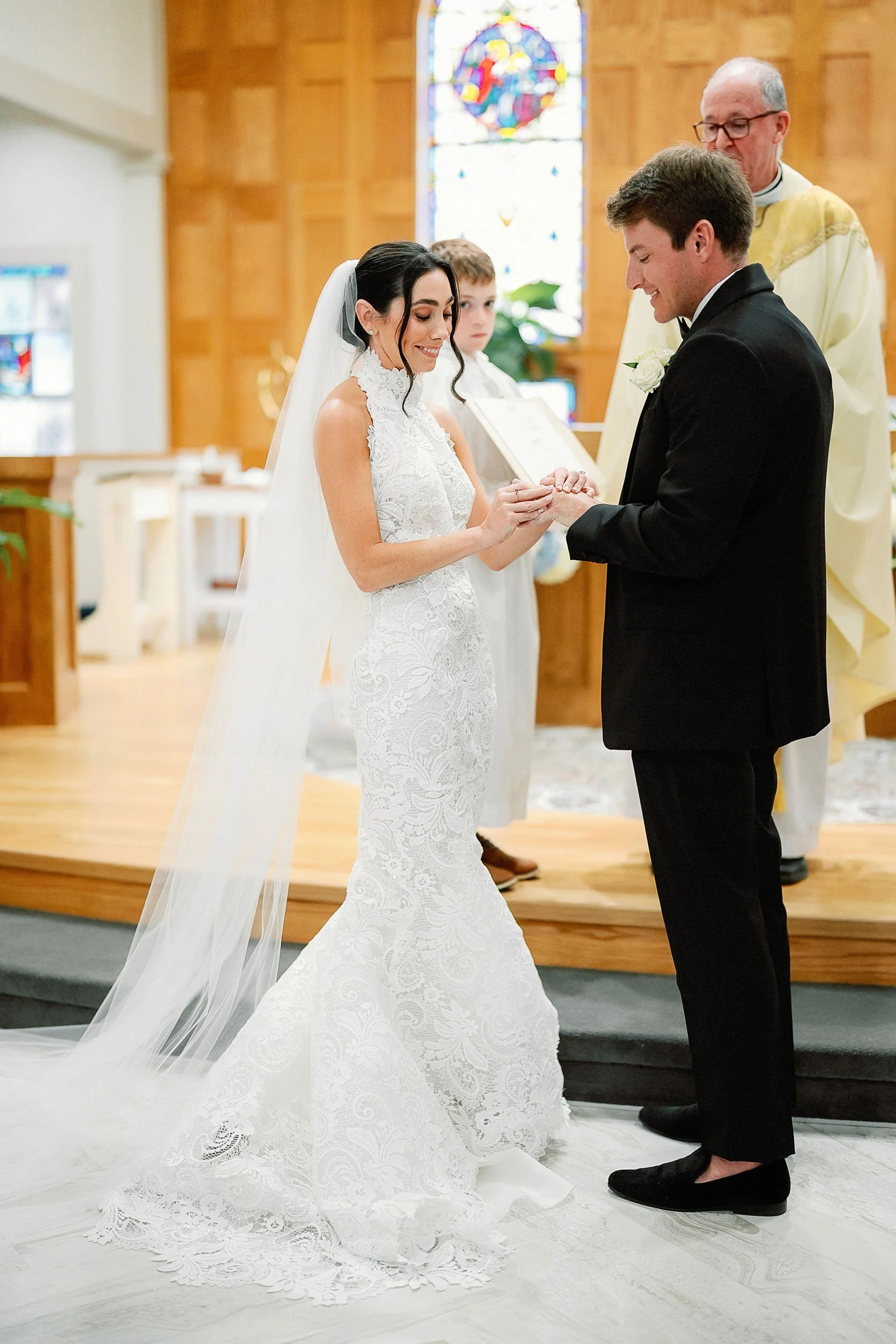 A bride and groom exchanging wedding rings during a ceremony in a church, with a priest standing behind them and a young boy observing.