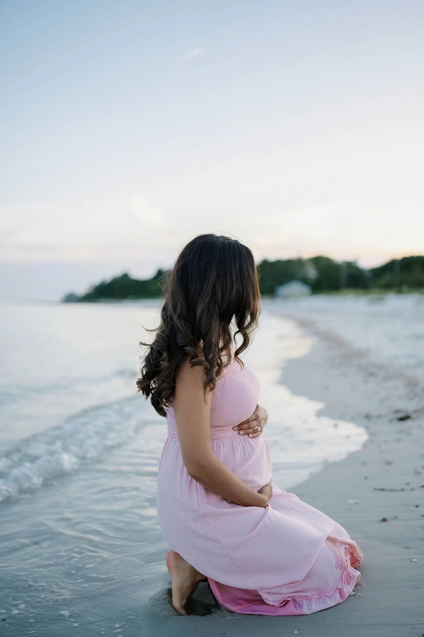 A pregnant woman with dark wavy hair wearing a pink dress kneeling on the beach near the shoreline, gently holding her belly with both hands.