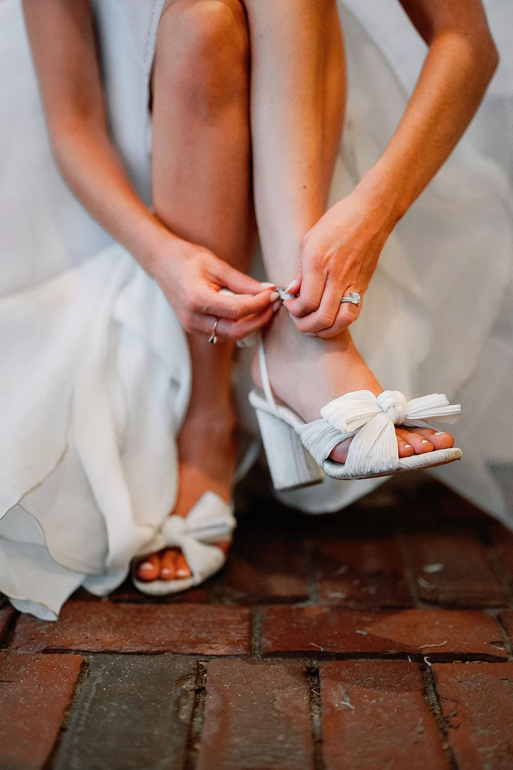 A bride is sitting and tying the strap of her white high-heeled sandal with a bow, wearing a wedding ring and a flowing white dress.