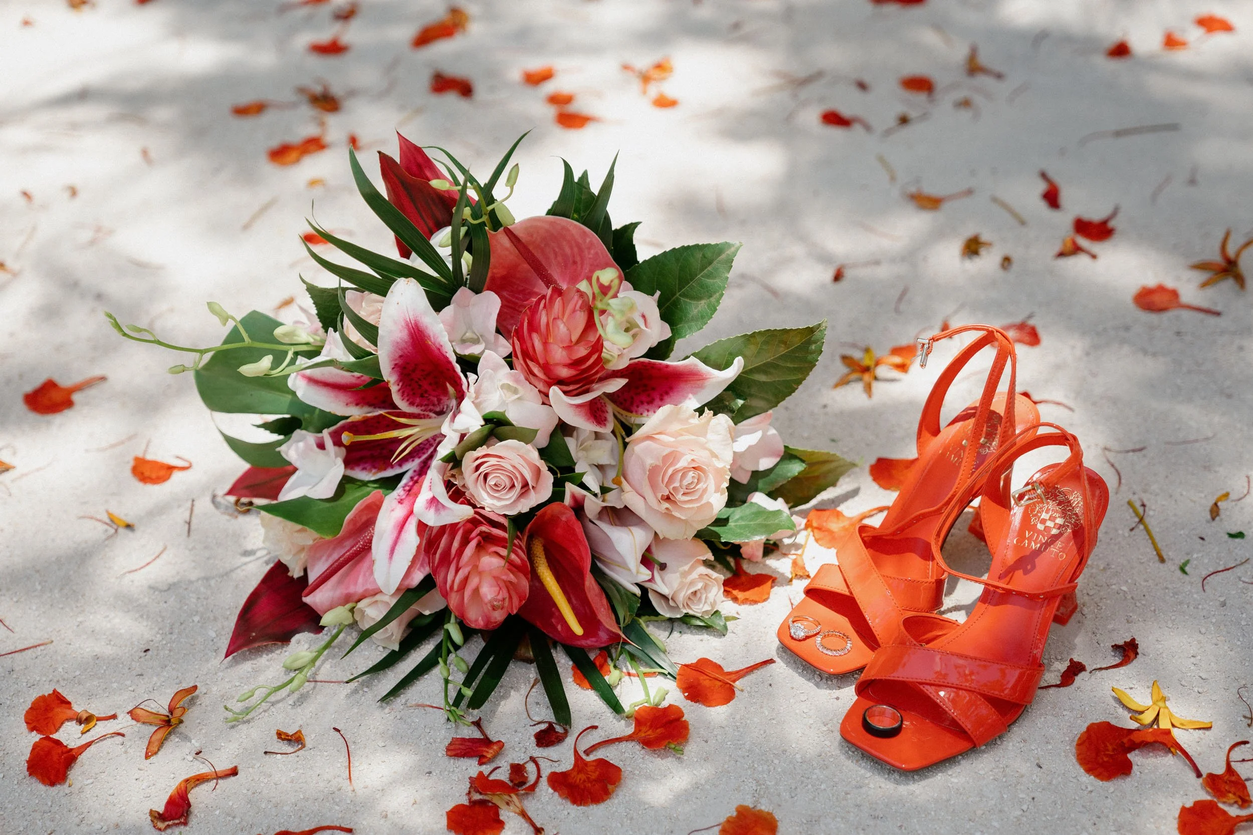 A bouquet of pink and white flowers and green leaves on sandy ground with scattered orange flower petals next to a pair of orange high-heeled sandals and rings.