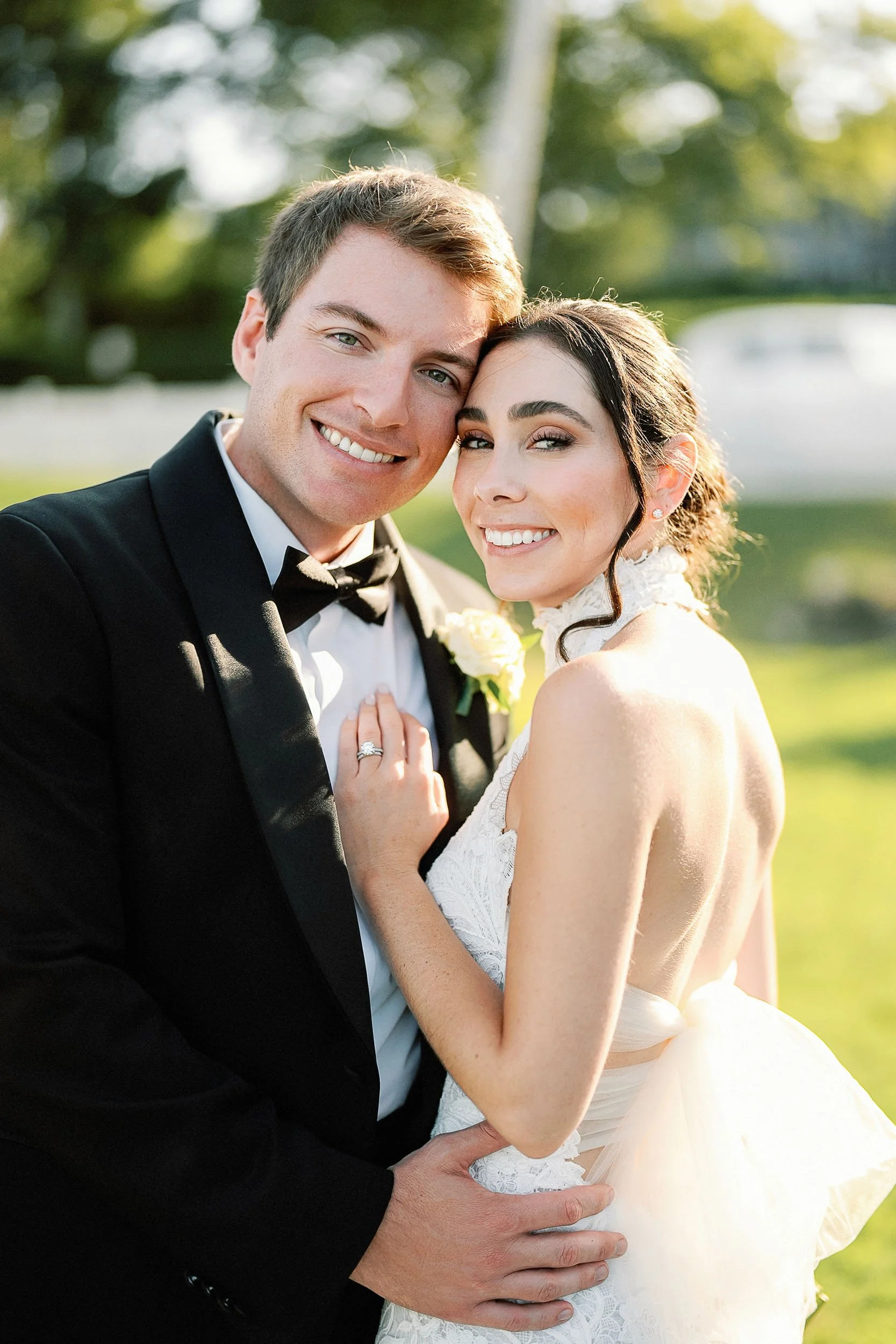 A happily smiling bride and groom embrace outdoors during their wedding, with sunlight filtering through trees in the background.