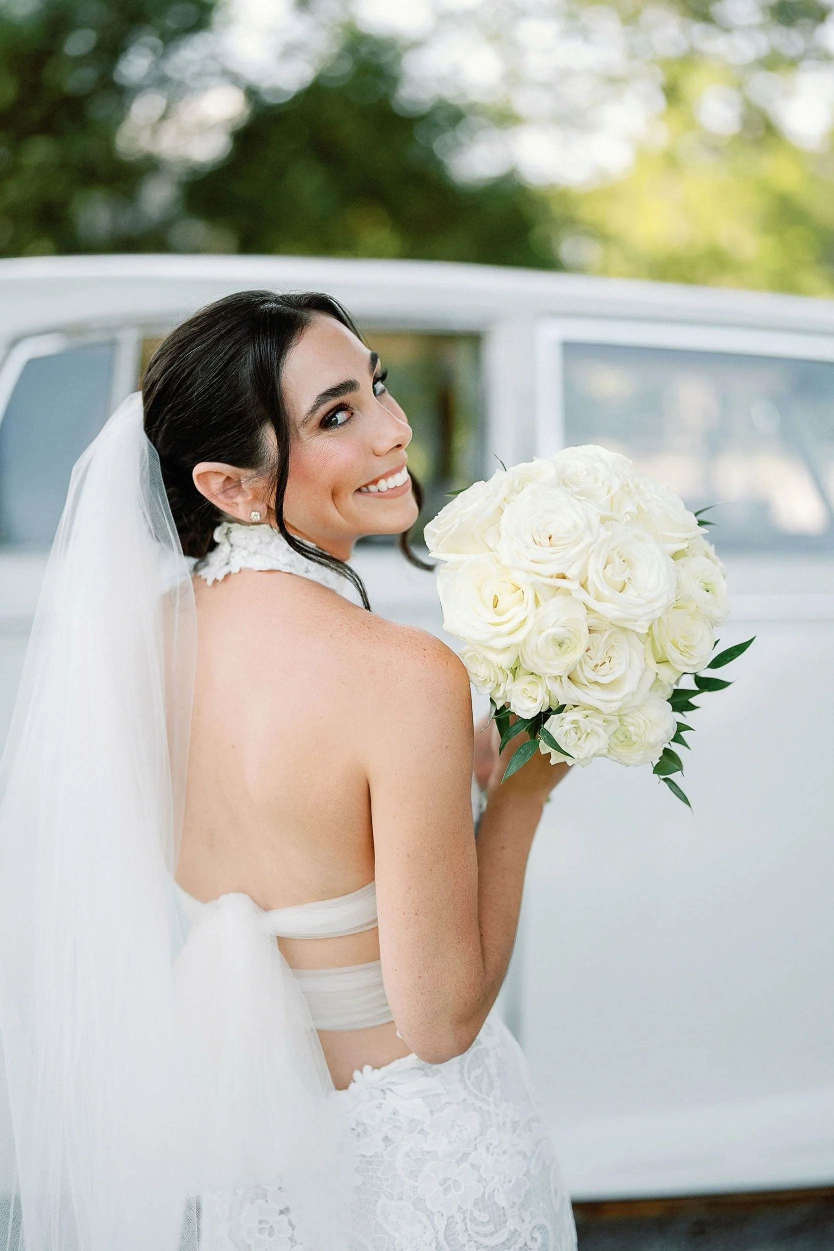 A bride with dark hair, smiling, holding a bouquet of white roses, standing outdoors with a vintage car in the background.