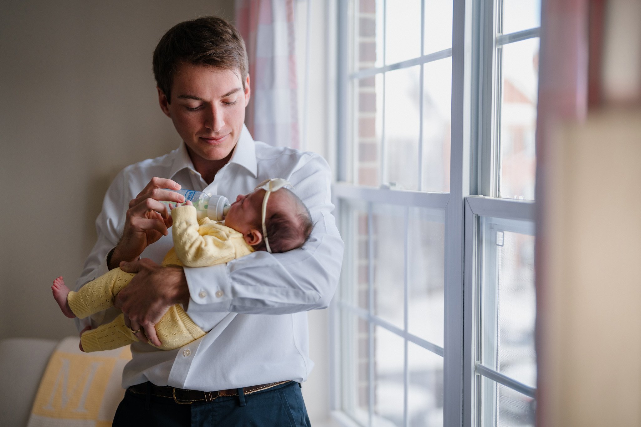 A man in a white shirt holding a baby with a feeding tube while feeding him with a bottle near a window.