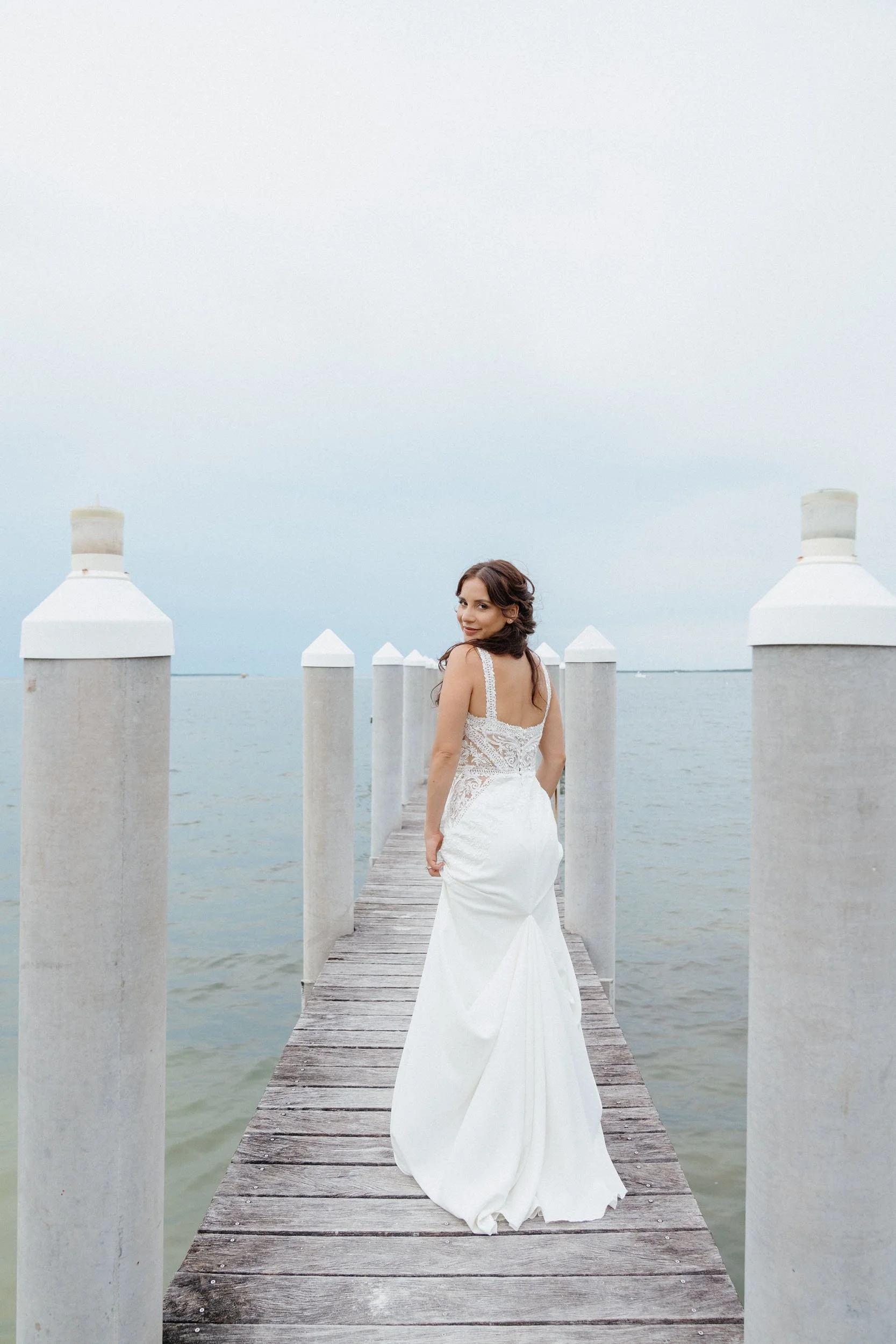 Woman in a white wedding dress standing on a wooden pier over water, looking back at the camera with overcast sky.