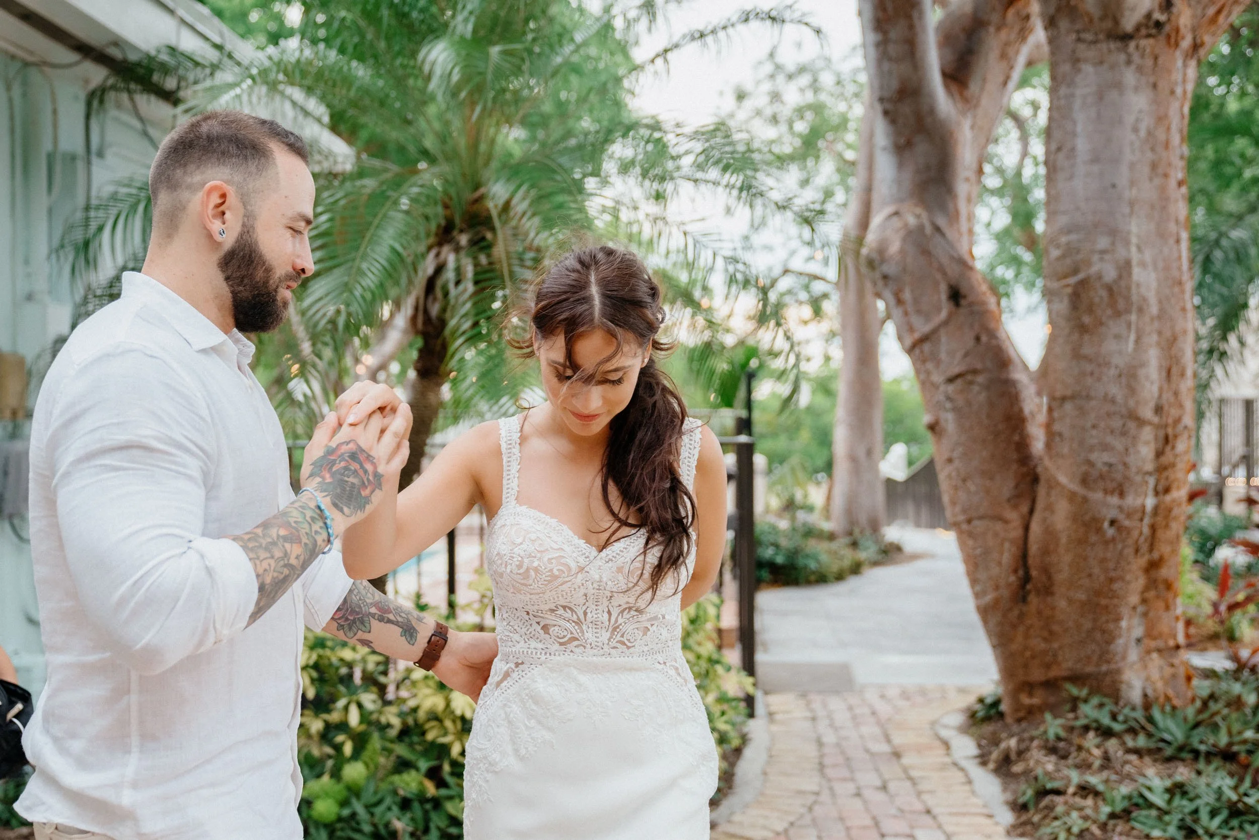 A couple holding hands and praying outdoors near trees and greenery, with the woman wearing a white lace dress.