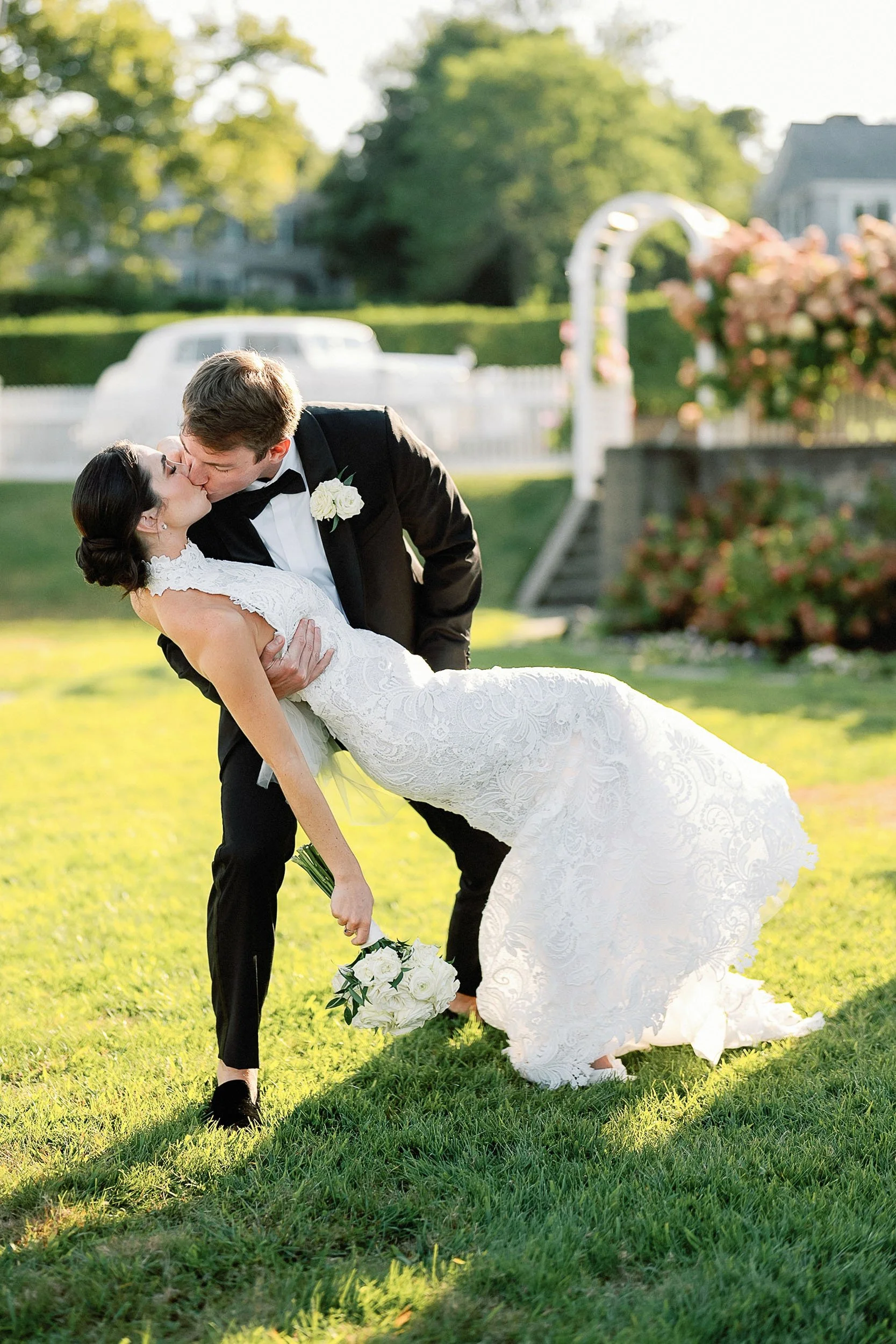 A newlywed couple sharing a kiss outdoors, with the groom dipping the bride in a grassy garden area during sunset.