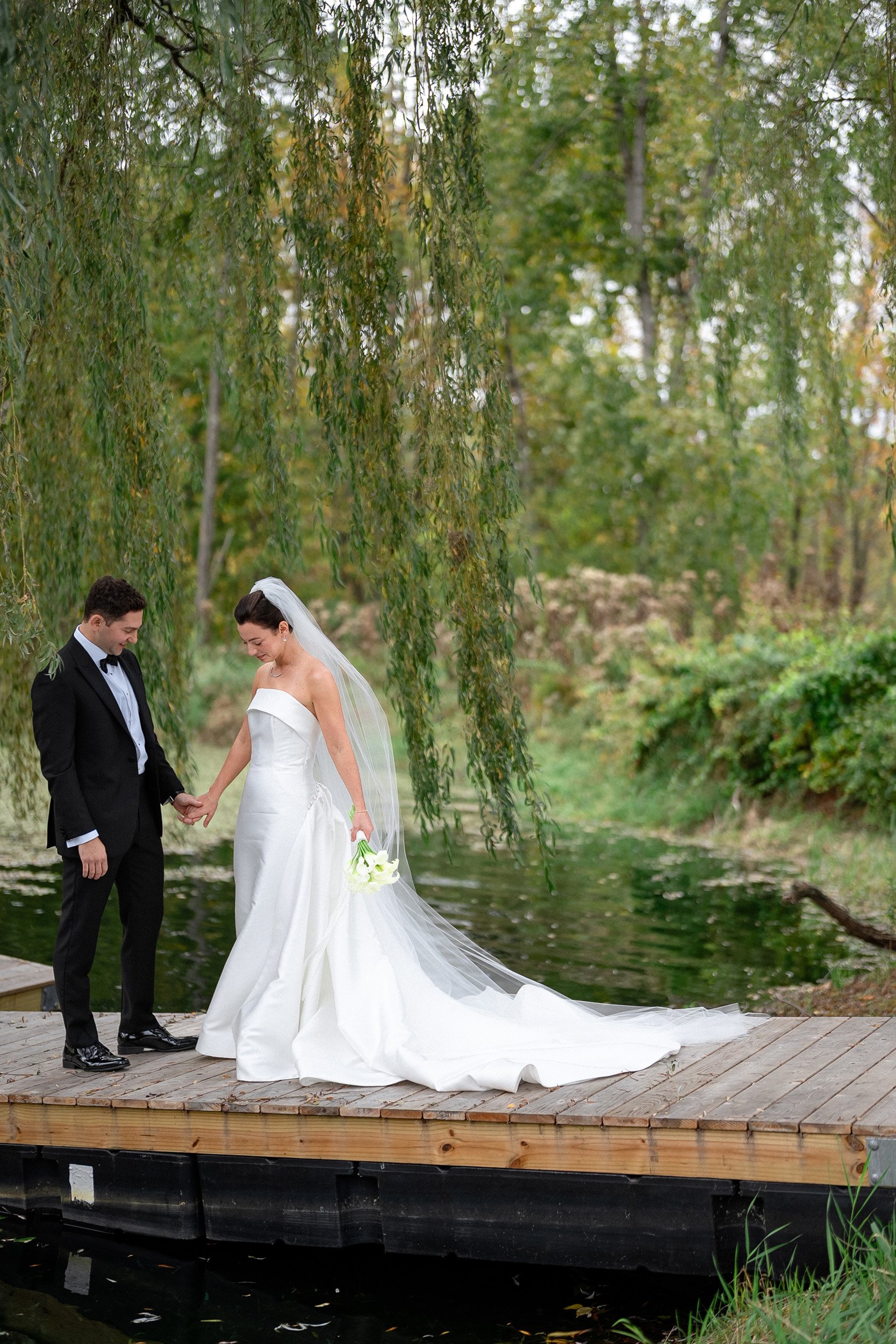A bride and groom holding hands on a wooden dock by a pond, surrounded by trees in a natural outdoor setting.