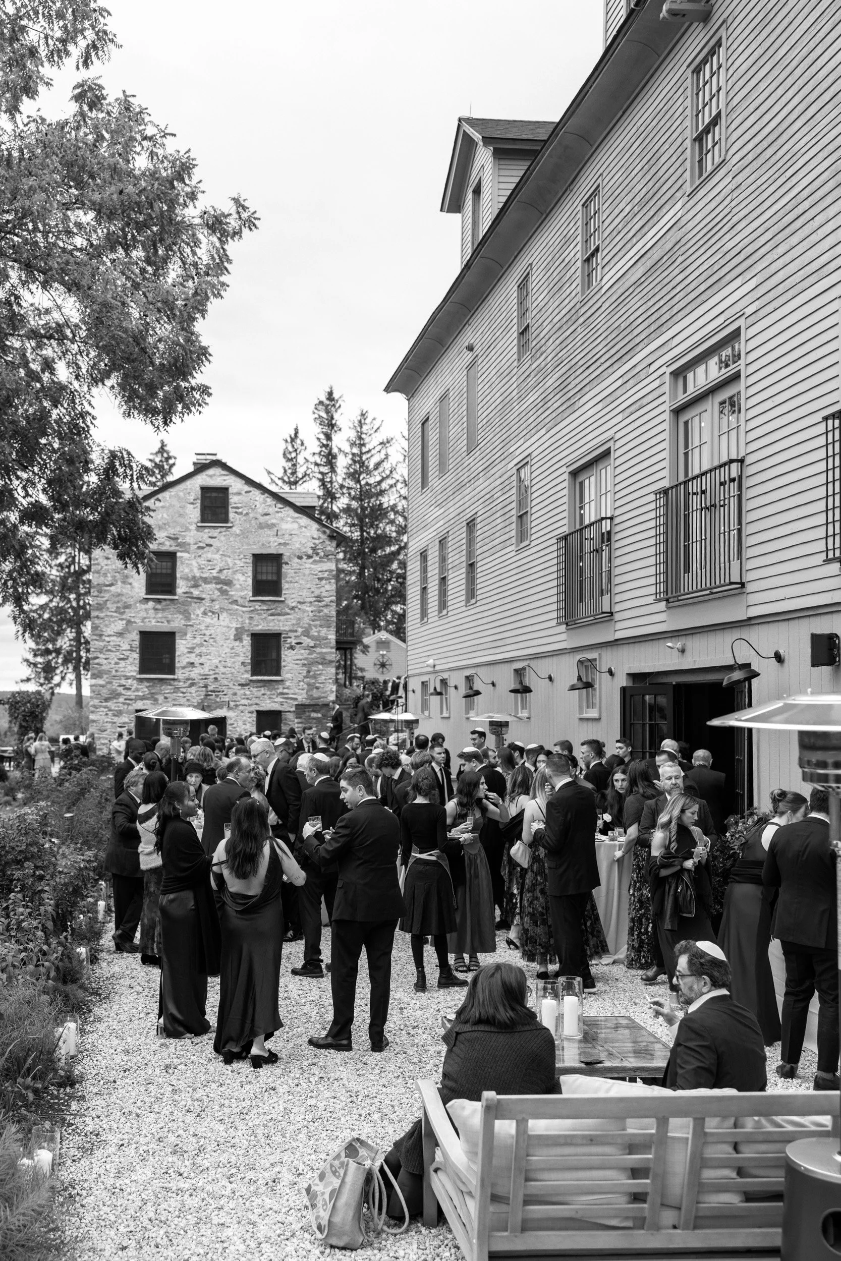 Black and white photo of a gathering of people in formal attire outside a building, with some standing and conversing and others sitting on a bench, with large windows, balconies, outdoor lamps, and trees in the background.