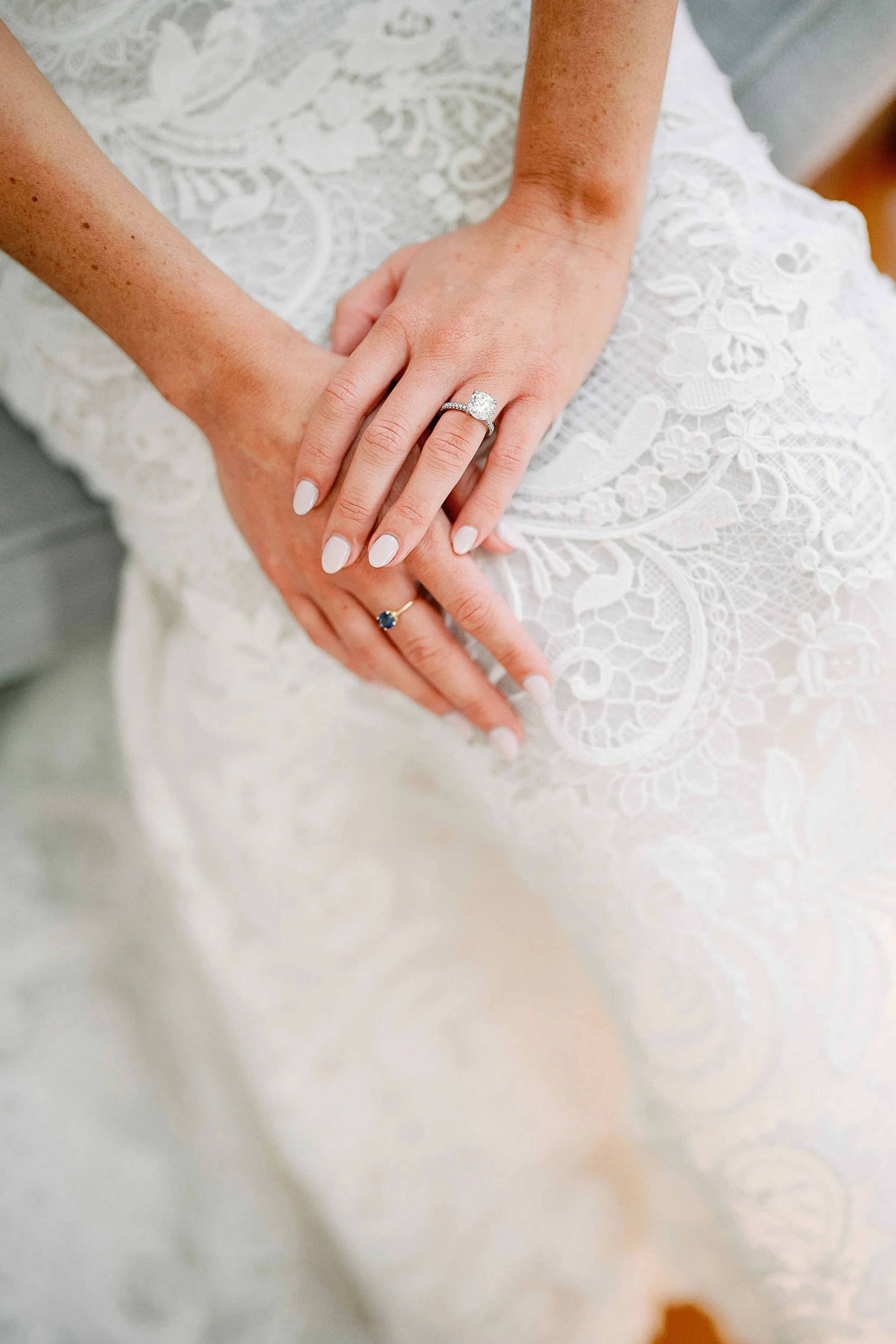 Close-up of a bride's hands with engagement ring and wedding band resting on her lap covered in white lace fabric.