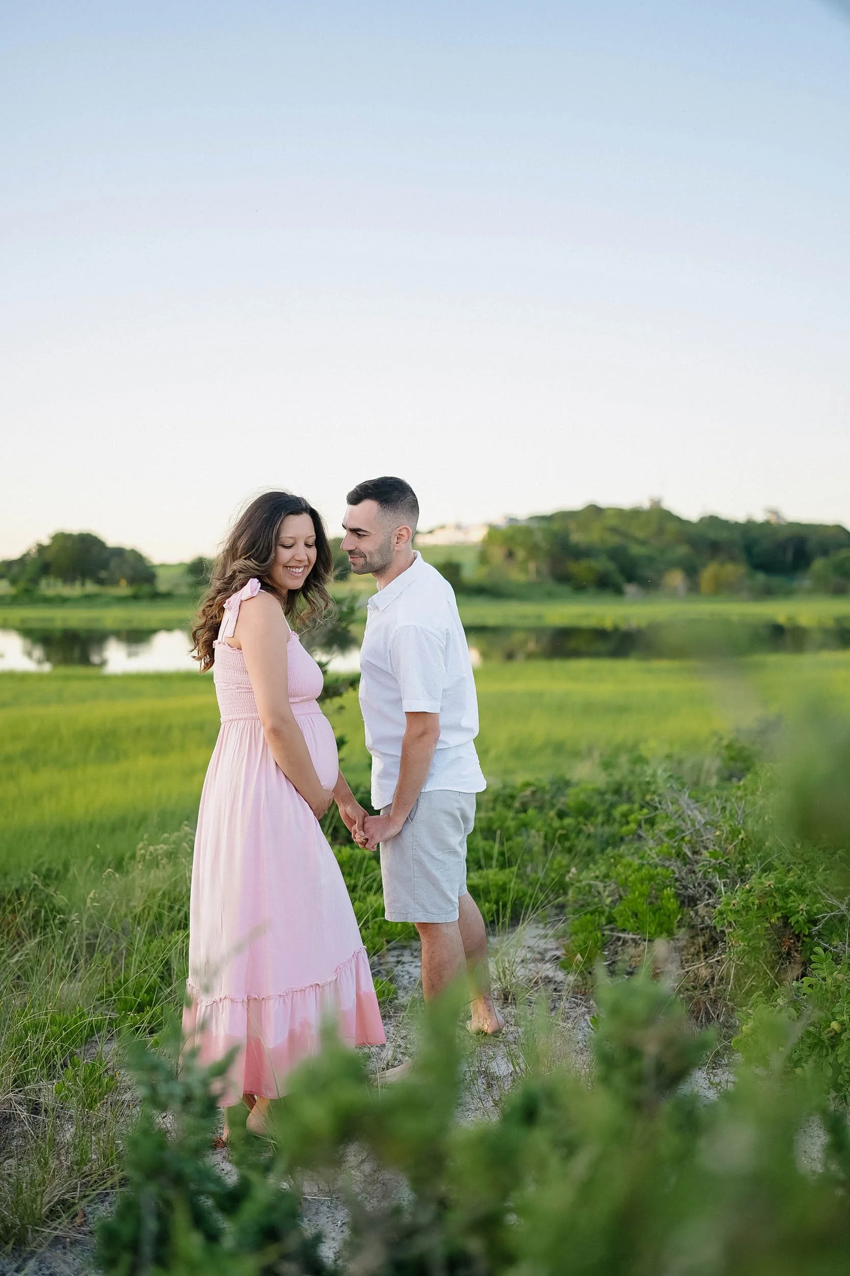 A pregnant woman and a man holding hands and smiling in a grassy outdoor setting with a pond and trees in the background.