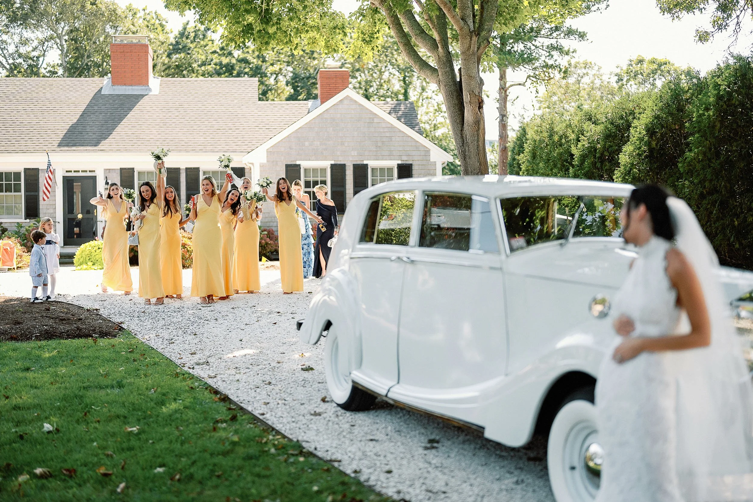 A bride stands in front of a vintage white car at an outdoor wedding. In the background, a group of bridesmaids in yellow dresses cheer and celebrate near a house with American flags and green trees.