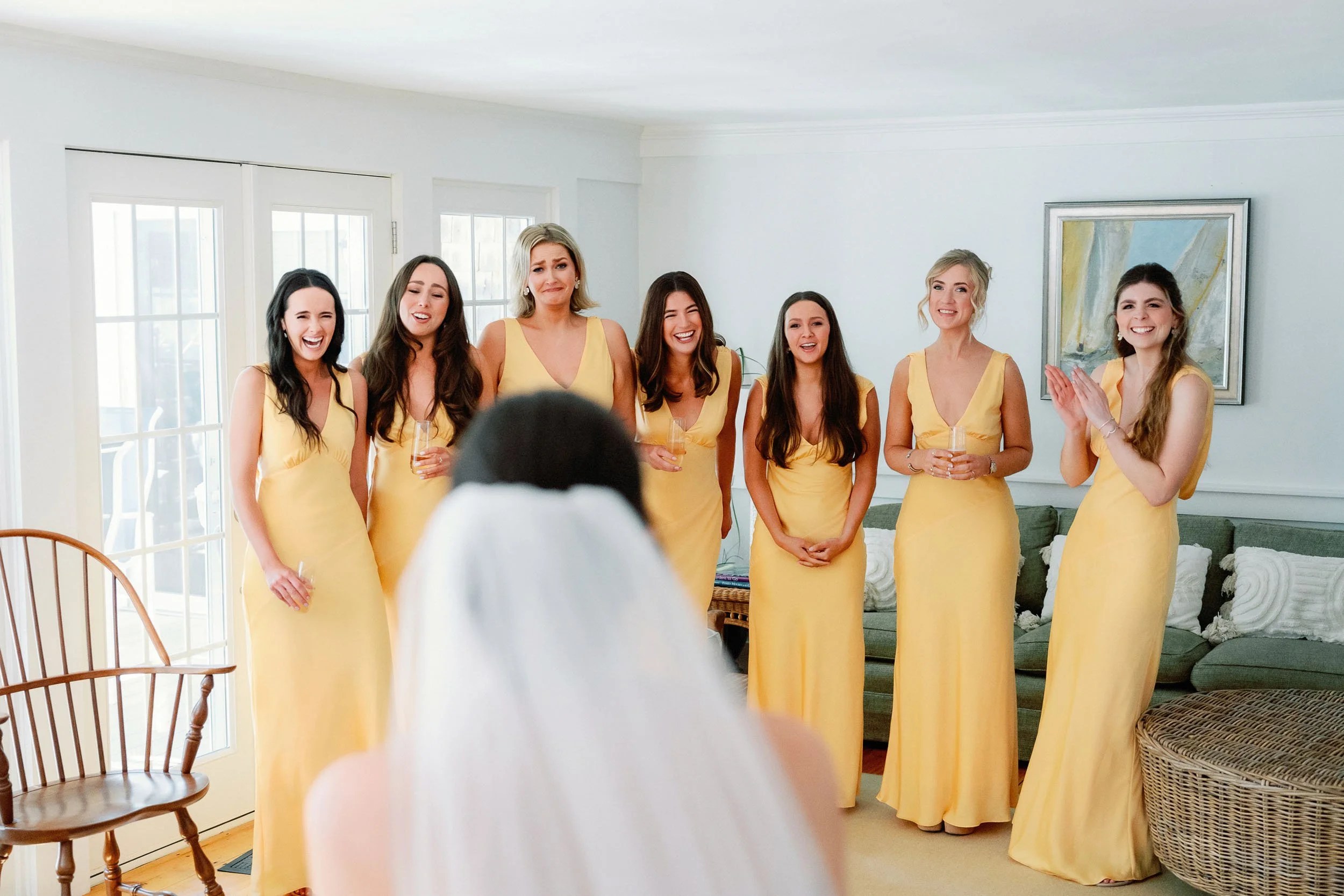 A bride with a white veil facing seven bridesmaids in yellow dresses, standing in a living room with a green sofa, wall art, and French doors.