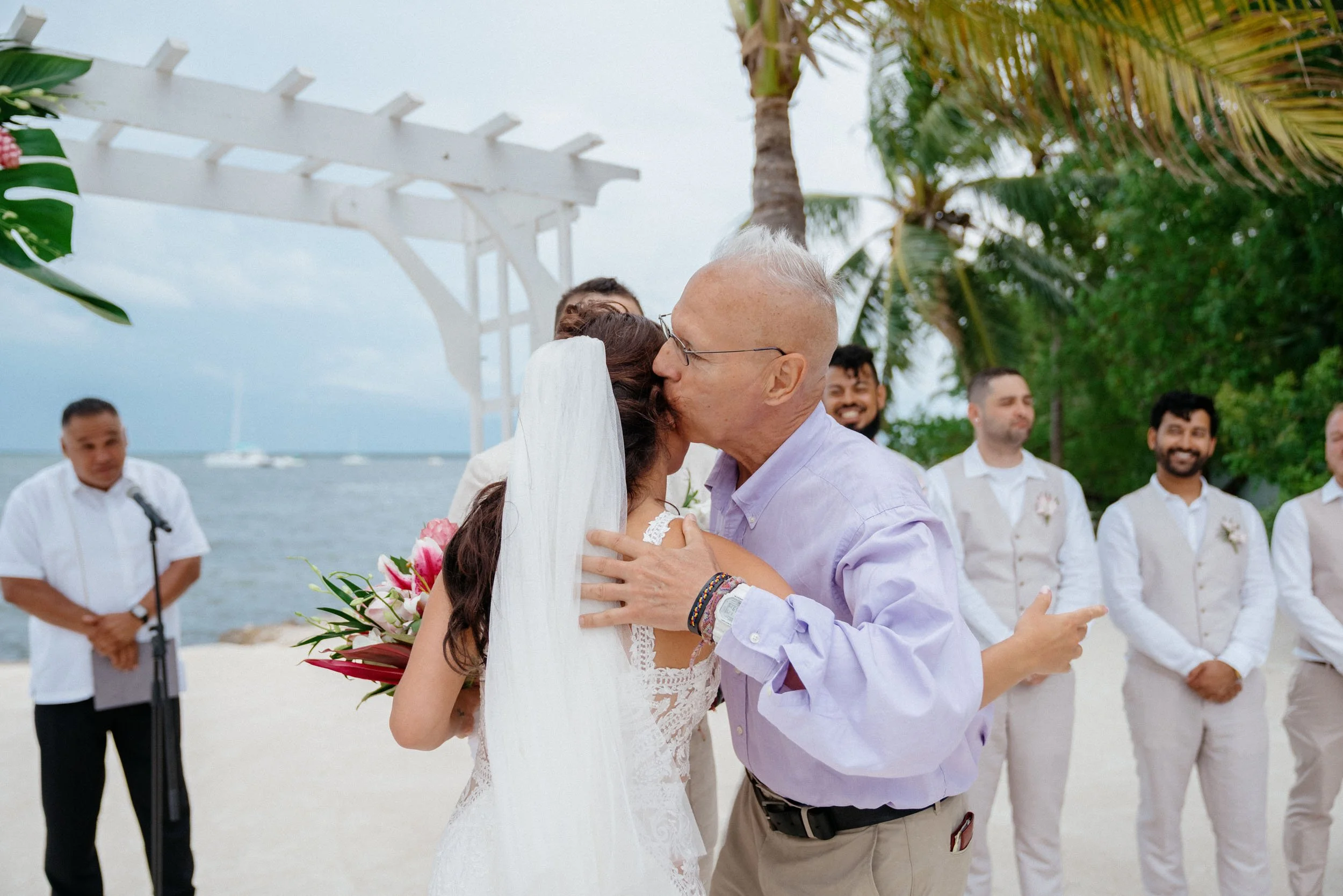 A man and woman kissing at a beach wedding ceremony, with several men smiling in the background and tropical trees nearby.