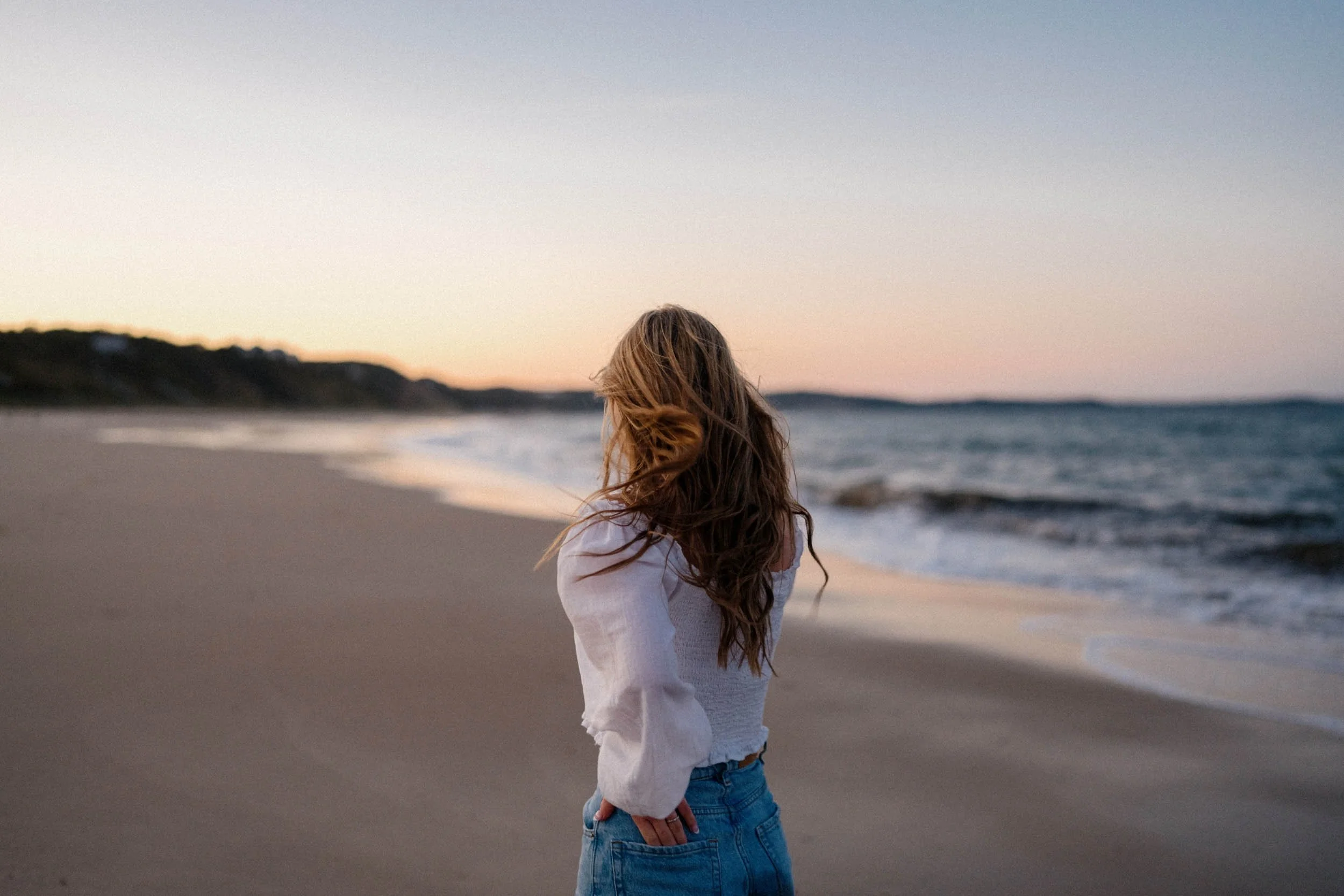 A woman with long hair stands on a beach at sunset, facing away from the camera towards the ocean.
