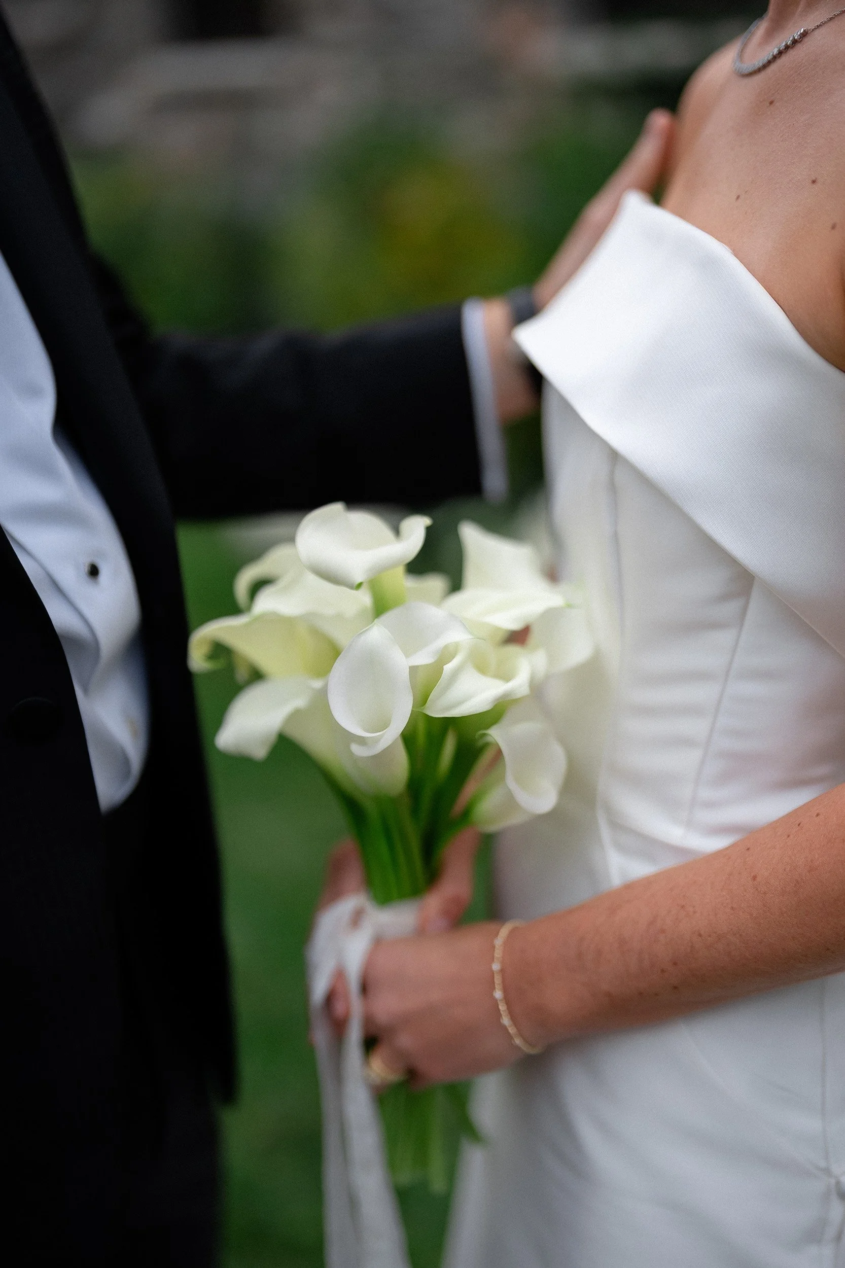 Bride holding a bouquet of white calla lilies during her wedding, with a groom partially visible in a tuxedo.
