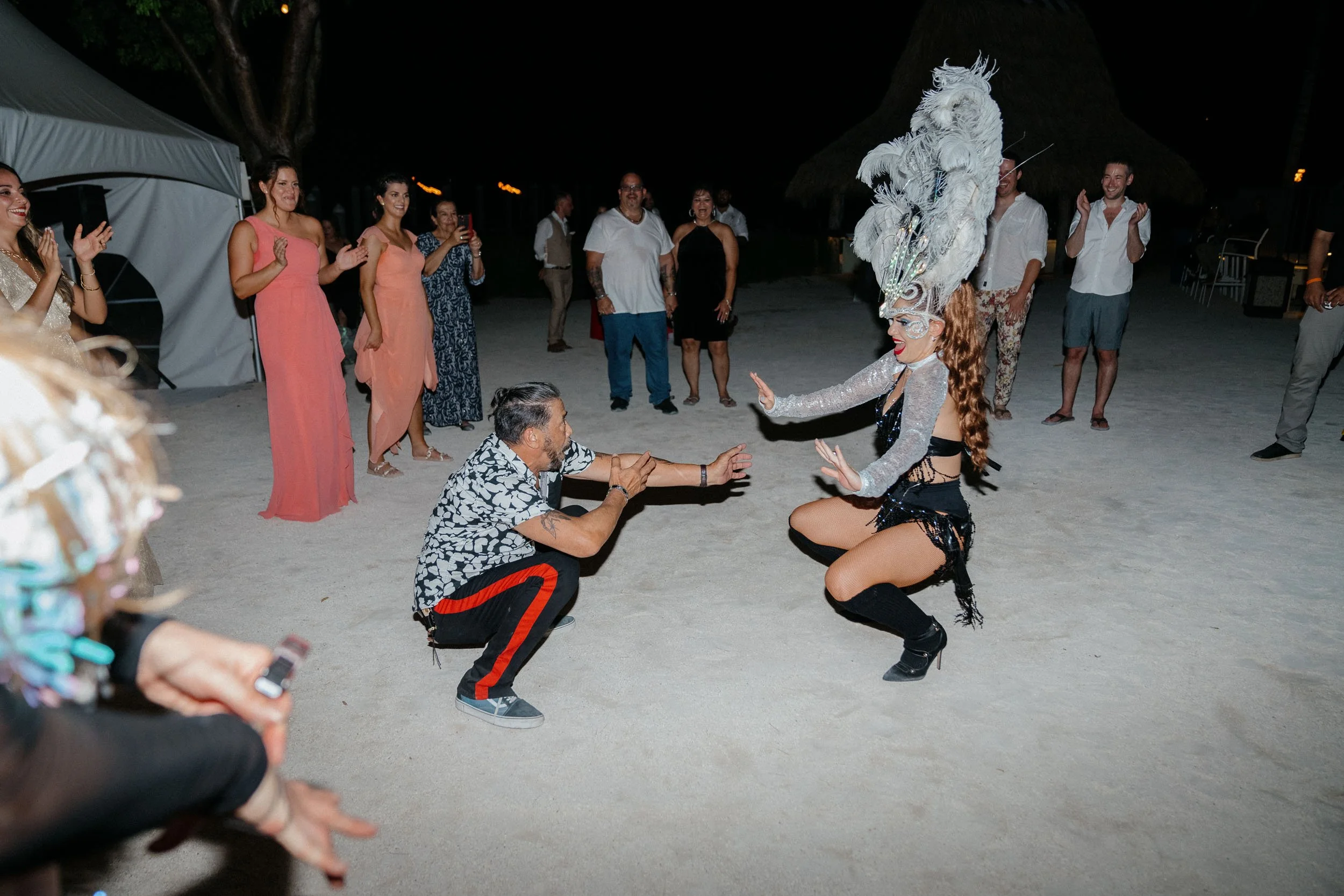 A woman in a showgirl costume with a large feather headdress performing a dance for a man squatting on the sand, surrounded by onlookers at an outdoor nighttime event.